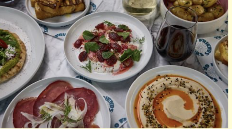 A variety of Mediterranean dishes on a table, including a plate of sliced cured meats, a bowl of roasted potatoes, a dish with hummus garnished with spices, a salad with greens and berries, a glass of red wine, and other plates with food.