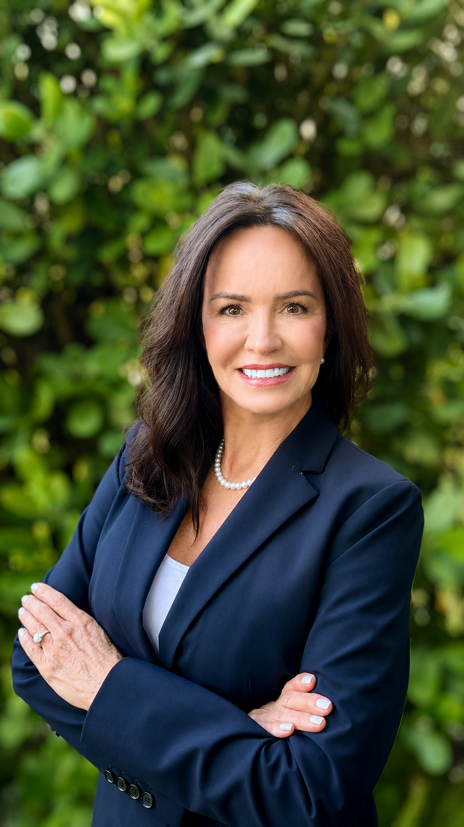 A smiling woman with dark brown hair wearing a navy blazer, white top, pearl necklace, and pearl earrings, standing outdoors with green foliage in the background.