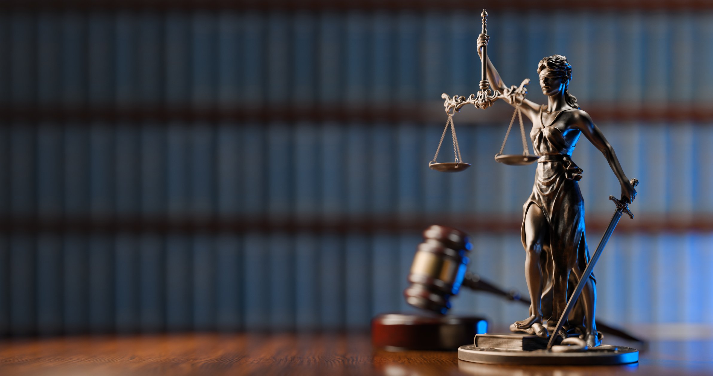 Bronze statue of Lady Justice holding scales and a sword, with a gavel on a wooden desk in the foreground, and a blurred bookshelf in the background.
