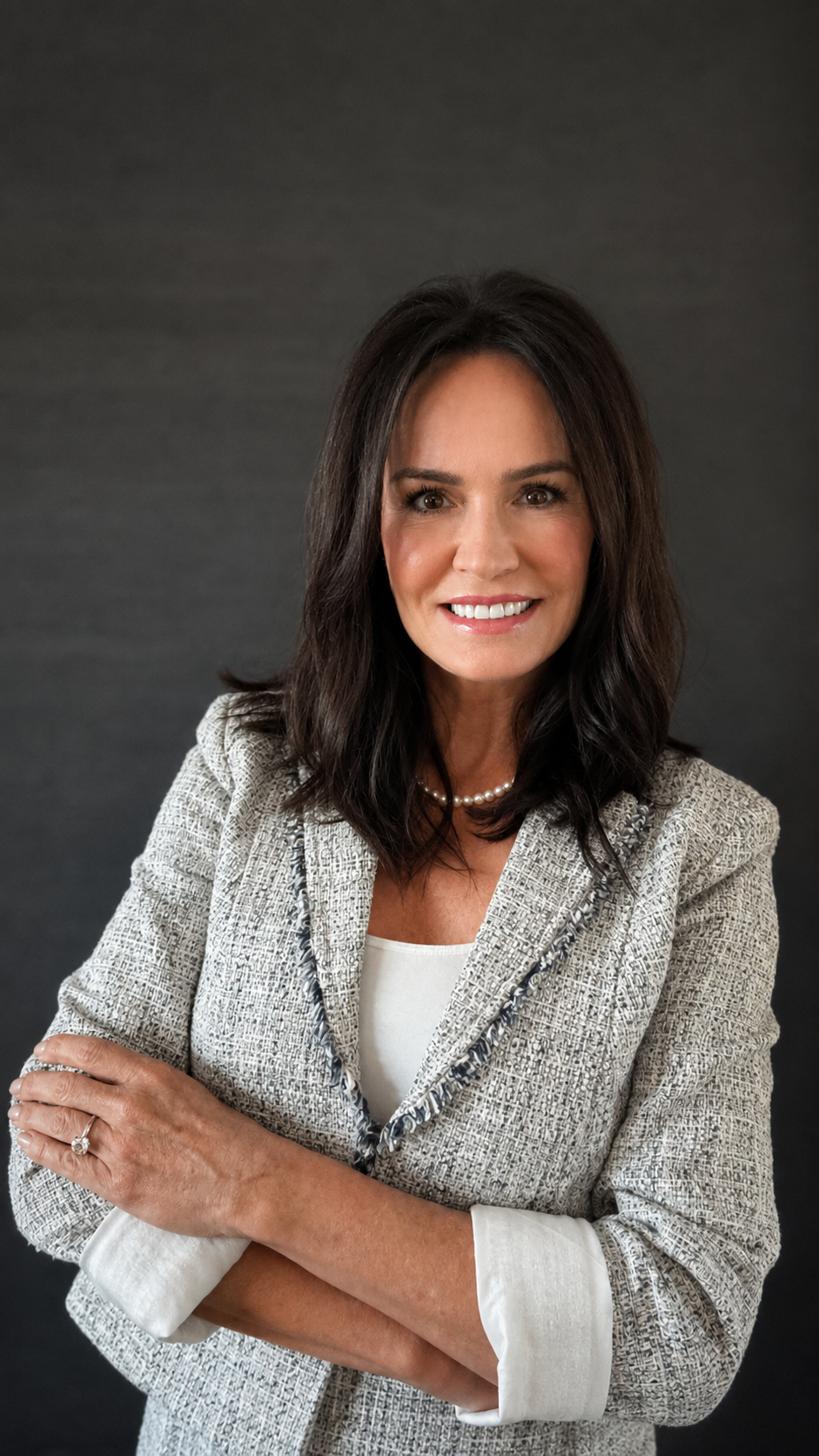 A woman with shoulder-length dark hair and a bright smile, wearing a light-colored textured blazer, a pearl necklace, and a white blouse, standing against a dark background.