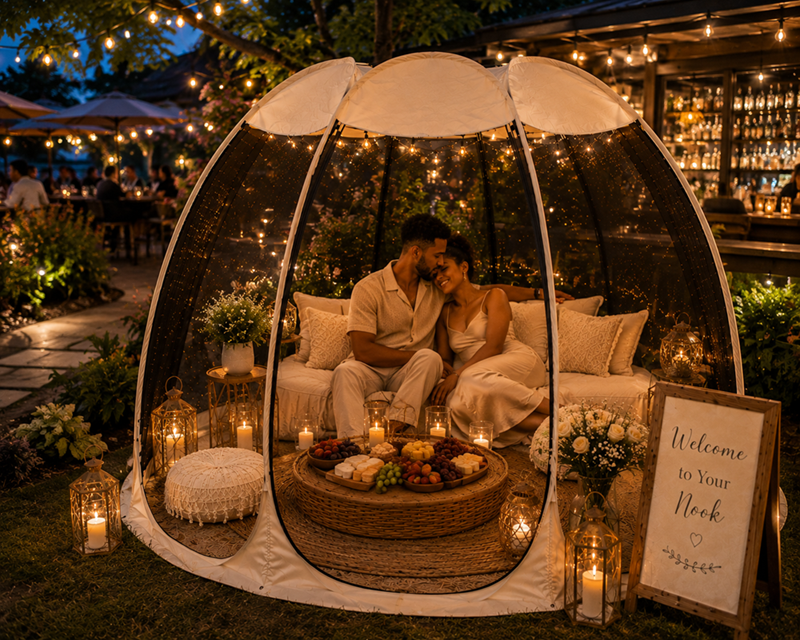 A couple sits inside a glowing, transparent outdoor igloo tent with string lights, surrounded by candles and floral decorations, at nighttime.