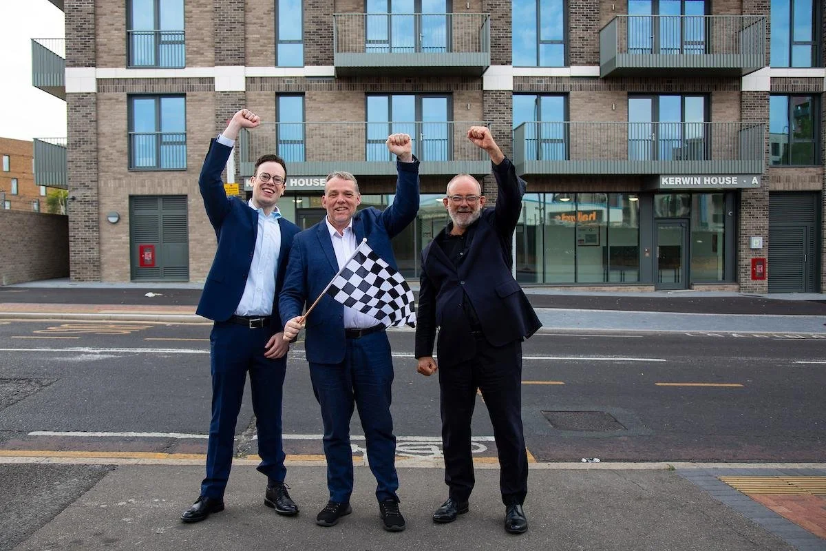 Three men in suits celebrating with raised fists, one holding a checkered flag, standing in front of a modern brick building with glass balconies.