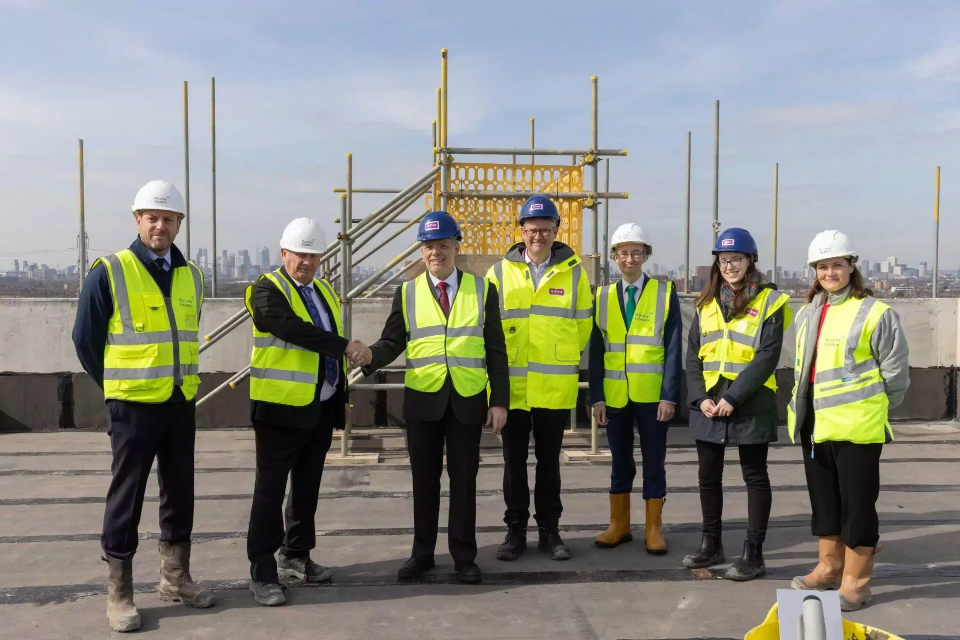 Group of seven construction professionals and executives standing on the roof of a building, wearing safety helmets and high-visibility vests, with a city skyline in the background. Two individuals are shaking hands, indicating a formal agreement or partnership.