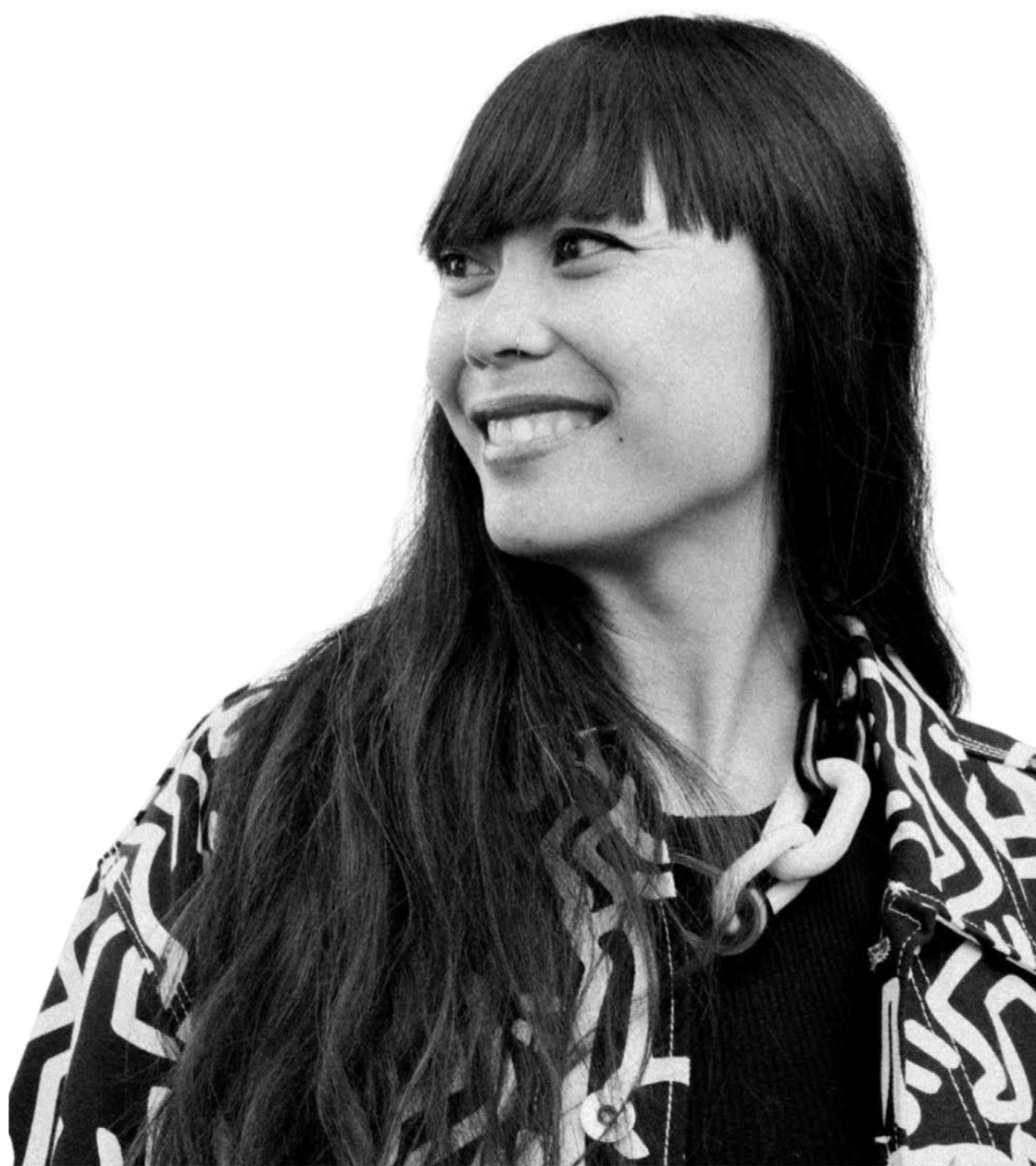 Black and white portrait of a woman with long hair and bangs, smiling and looking to the side, wearing a patterned jacket