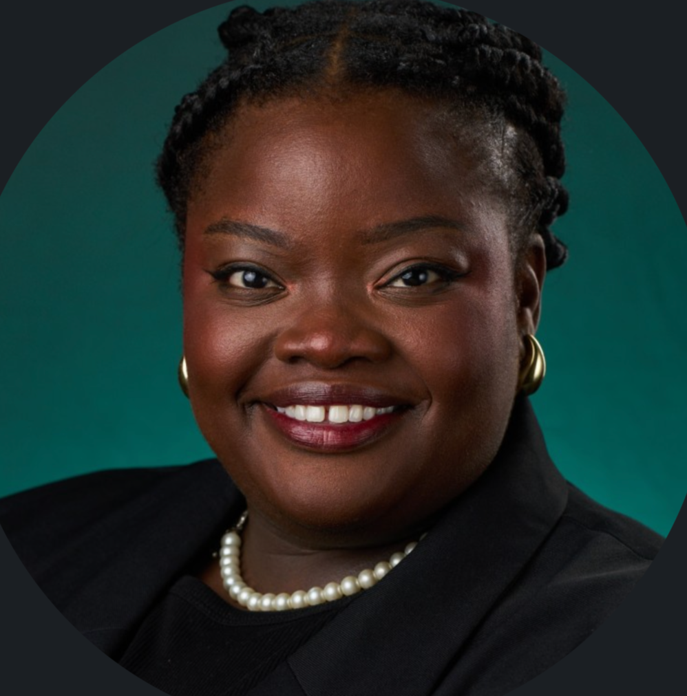 Portrait of a smiling Black woman wearing a black blazer, a pearl necklace, and pearl earrings, against a dark green background.