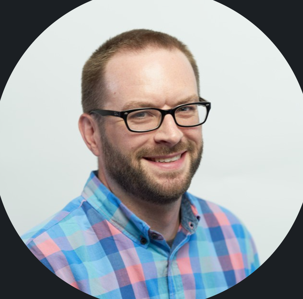 Headshot of a man with short brown hair, beard, glasses, and a bright smile, wearing a colorful plaid shirt.