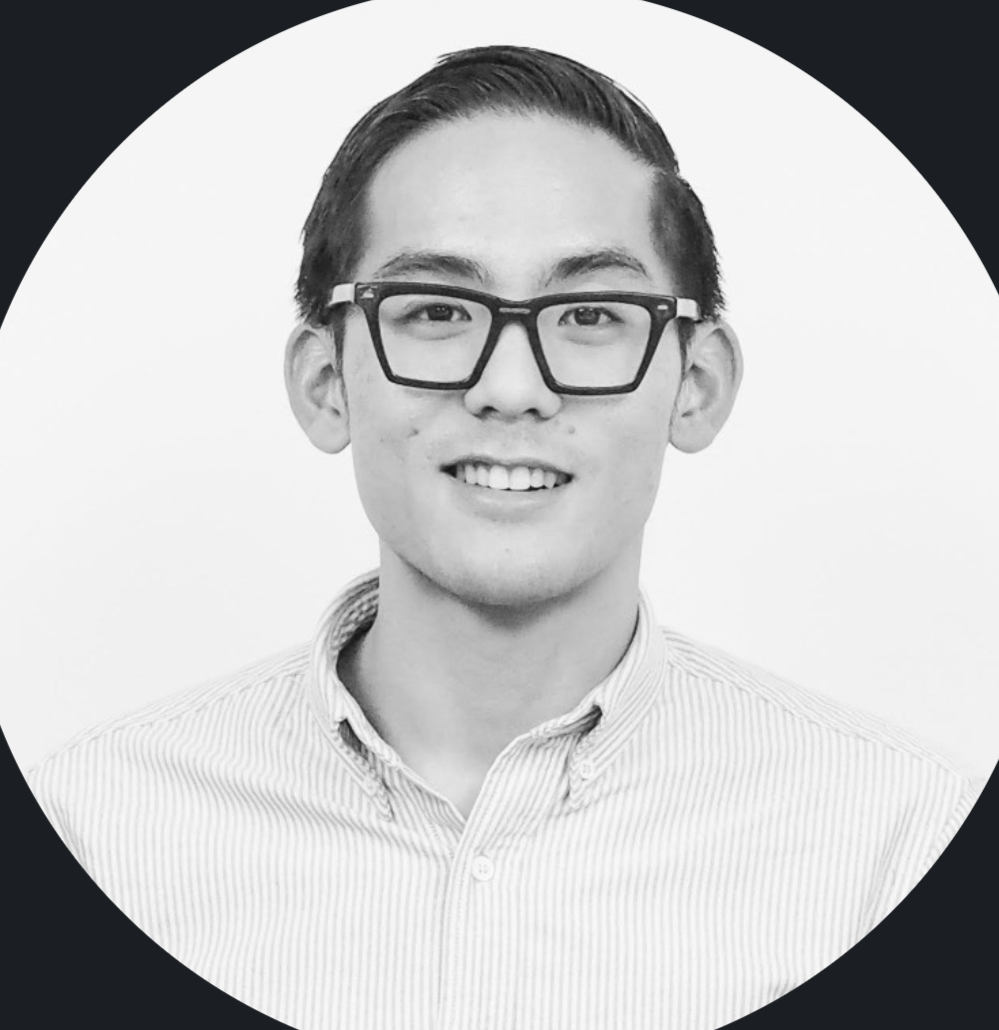 A black and white portrait of a young man with glasses, smiling, wearing a collared shirt.
