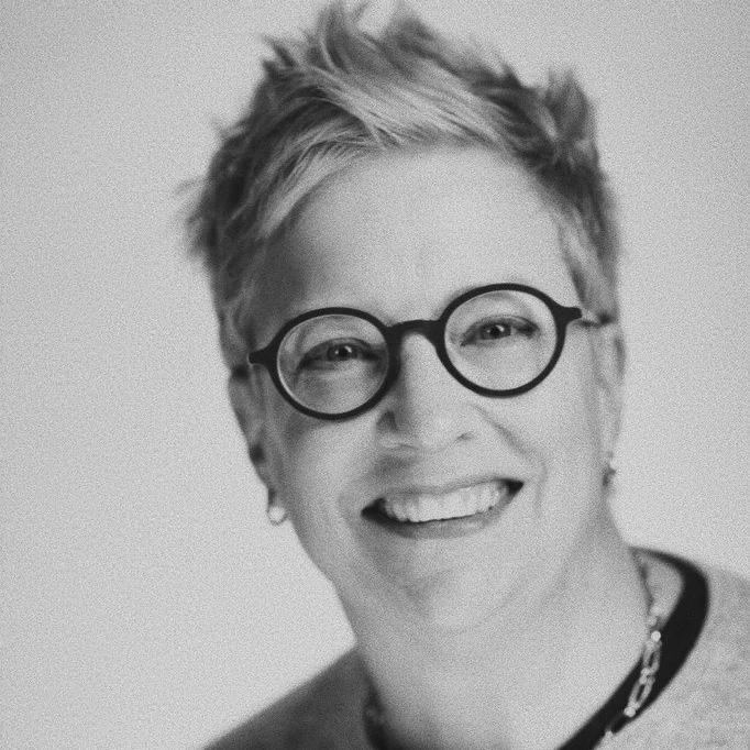 A smiling woman with short, spiky hair, round glasses, and earrings. She is wearing a necklace and a top with a dark collar.