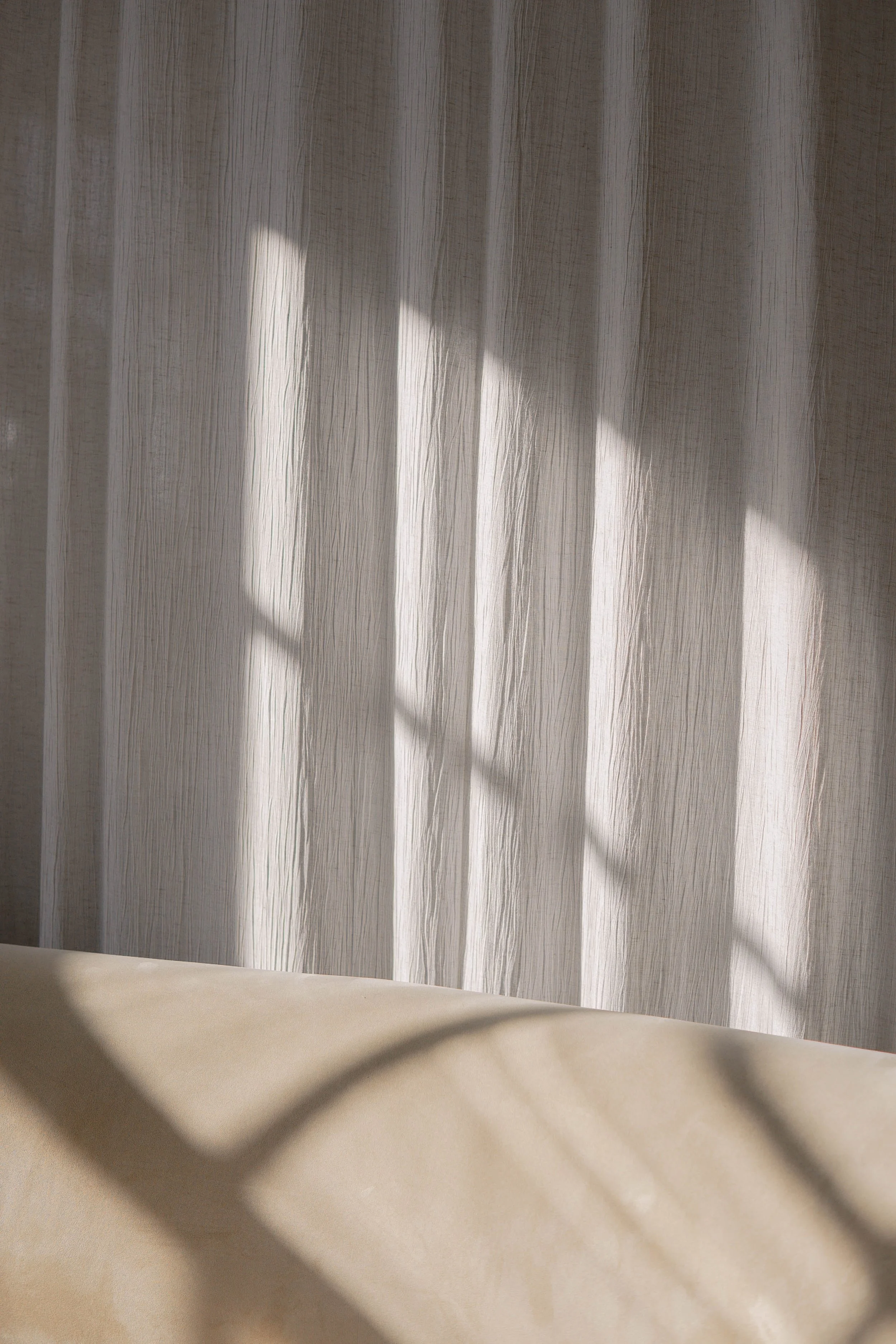 Light-colored curtain with shadows cast on it through a window, with a beige fabric surface in the foreground.