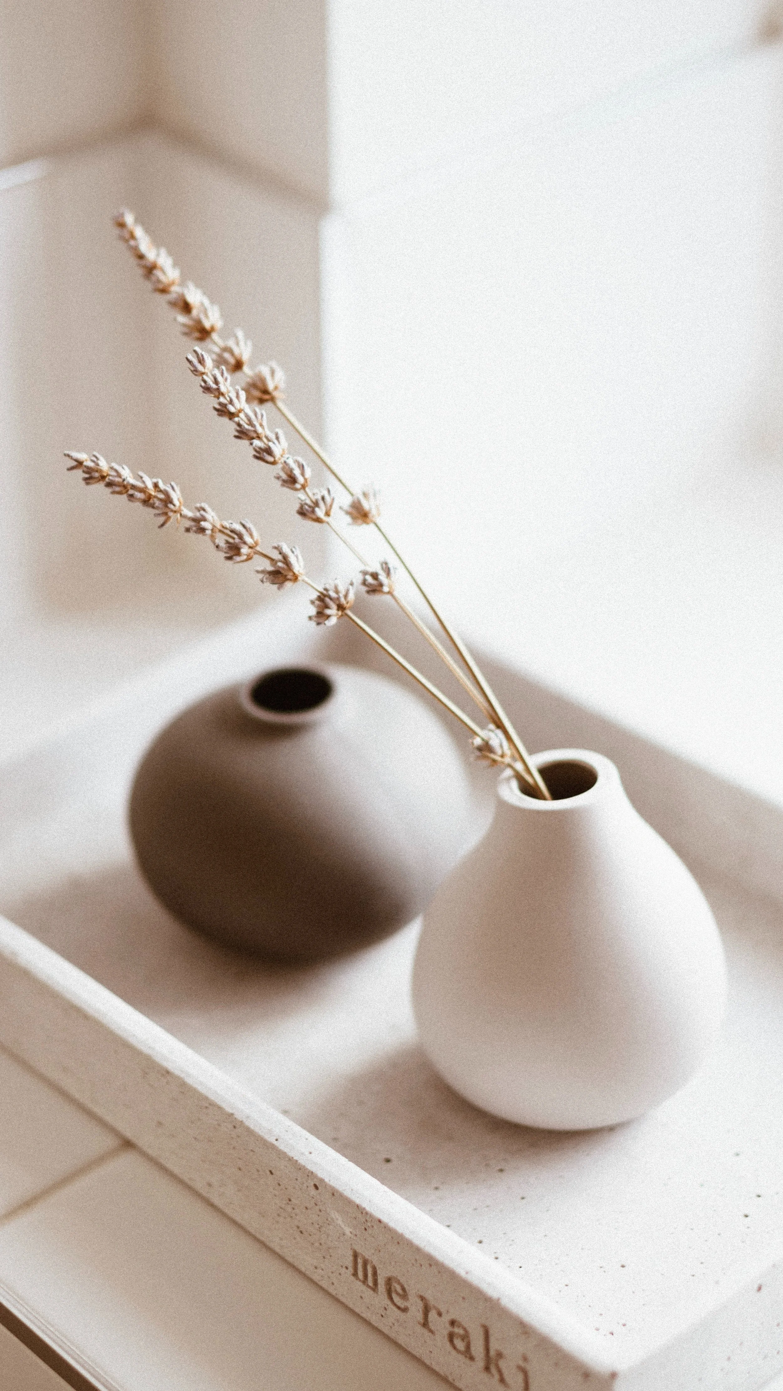 Two minimalist ceramic vases, one black and one white, with dried lavender in the white vase, placed on a white tray with the word 'Deraki' engraved on the side, indoors with soft natural lighting.