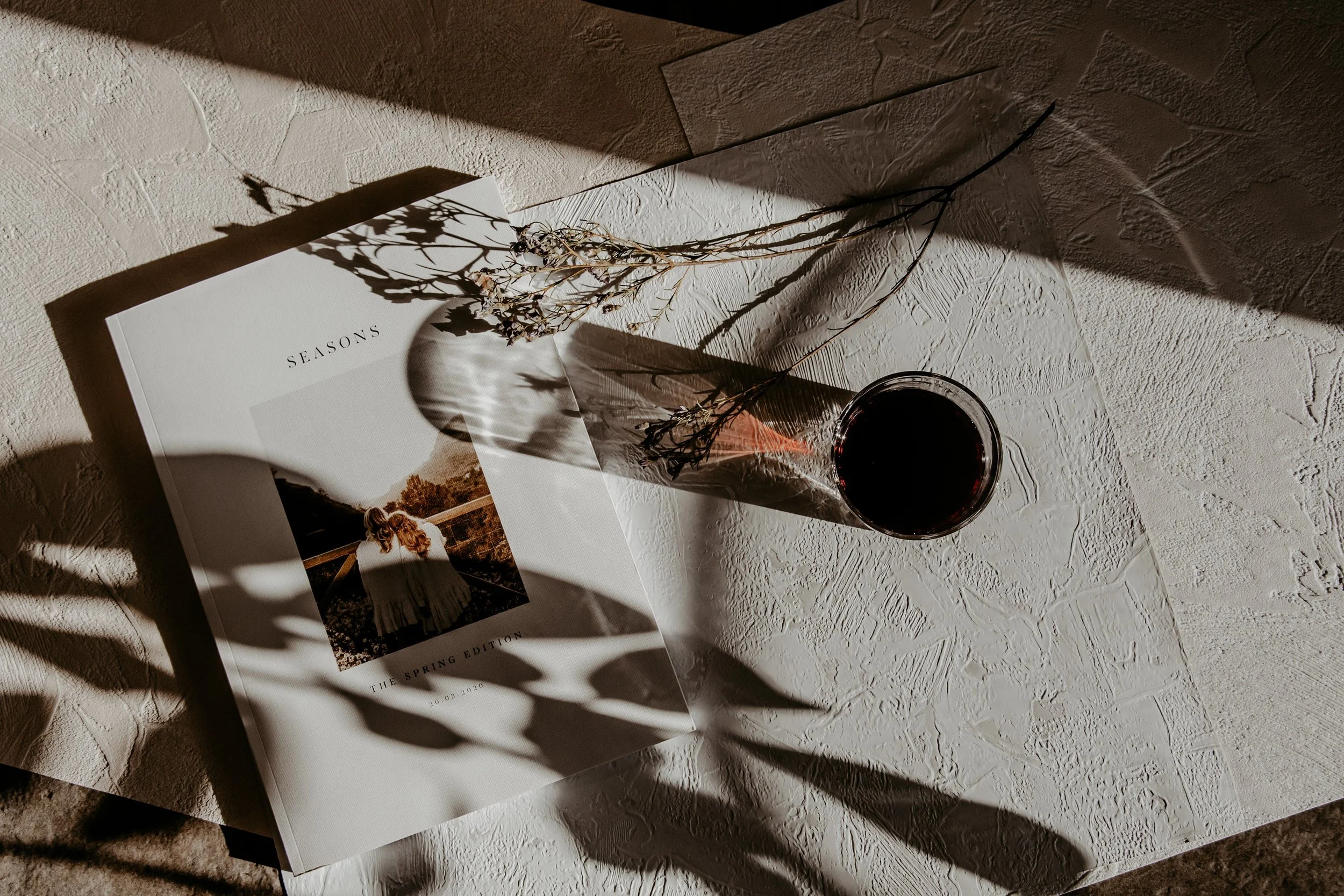 A magazine labeled 'SEASONS' is on a textured surface, with dried flowers and a glass of red wine casting shadows on the page.