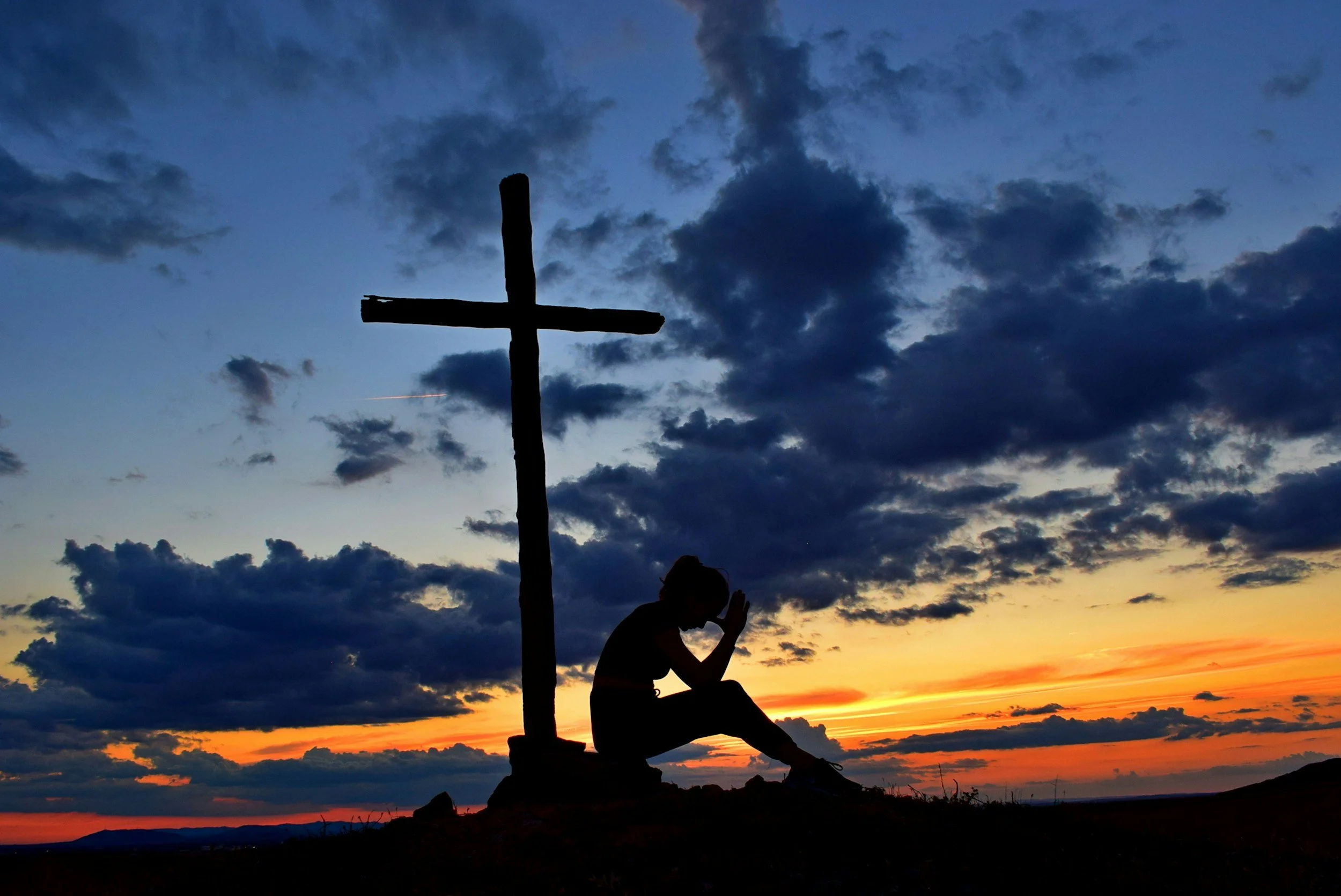 Silhouette of a person praying or meditating next to a wooden cross during sunset, with a colorful sky and scattered clouds.