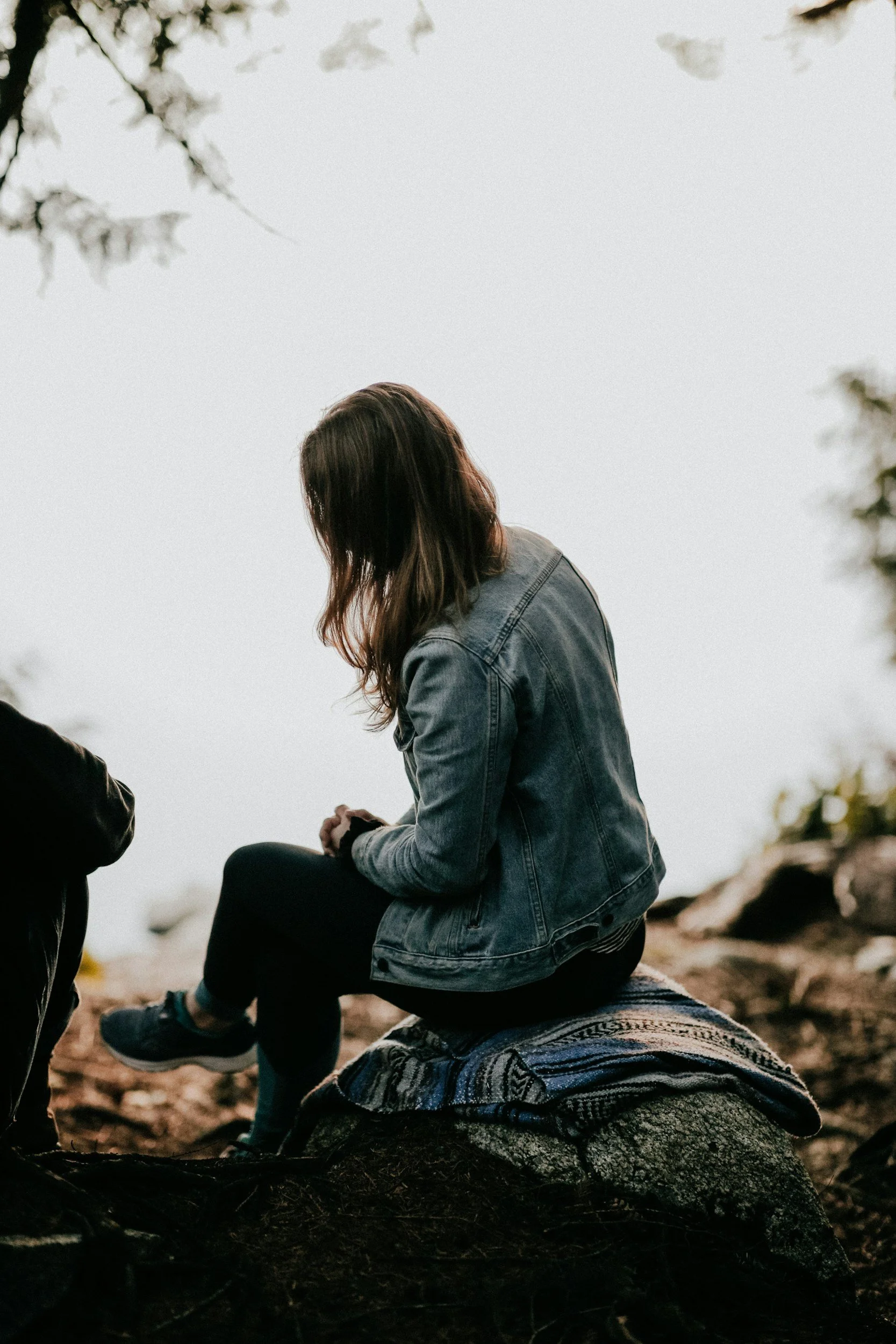 A person with long hair wearing a denim jacket is sitting outdoor on a rock, with their head bowed.