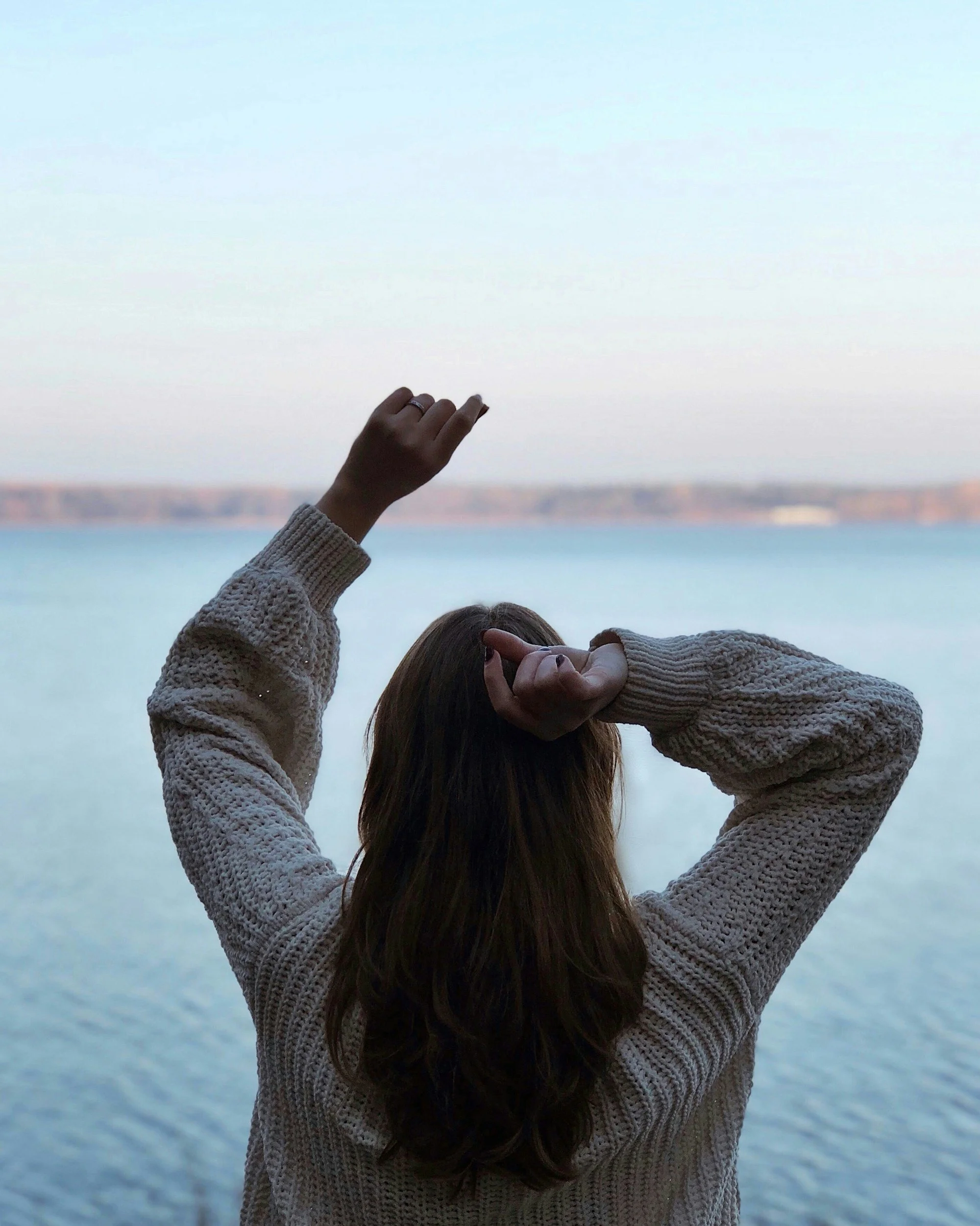 A person with long hair wearing a beige sweater, raising one arm and making a peace sign with the other, standing in front of a body of water with a blurred shoreline in the background.