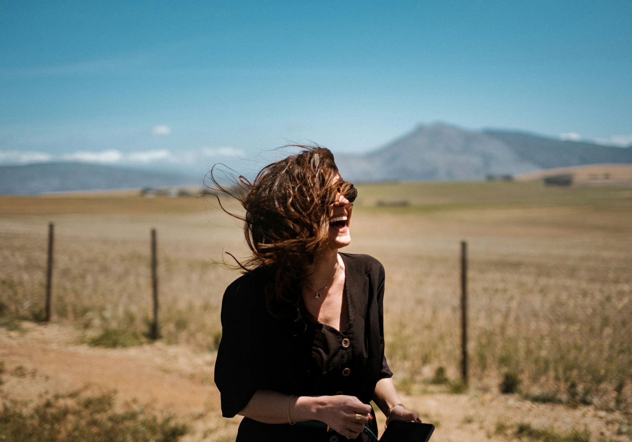 A woman with long, wavy hair smiling outdoors on a sunny day, holding a smartphone, with mountains and a clear blue sky in the background.