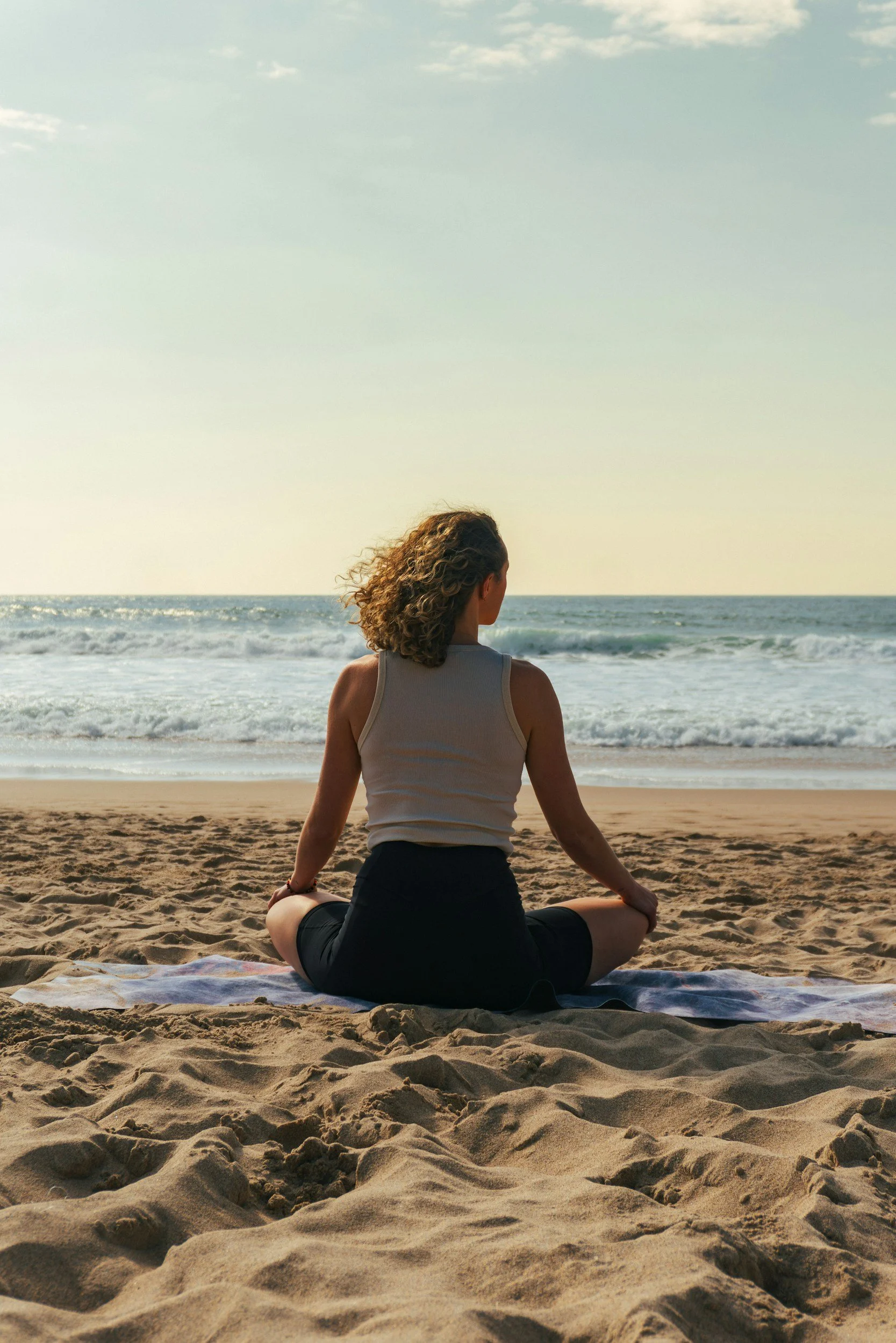 A woman practicing yoga on the sandy beach, sitting cross-legged in meditative pose facing the ocean during sunset or sunrise.