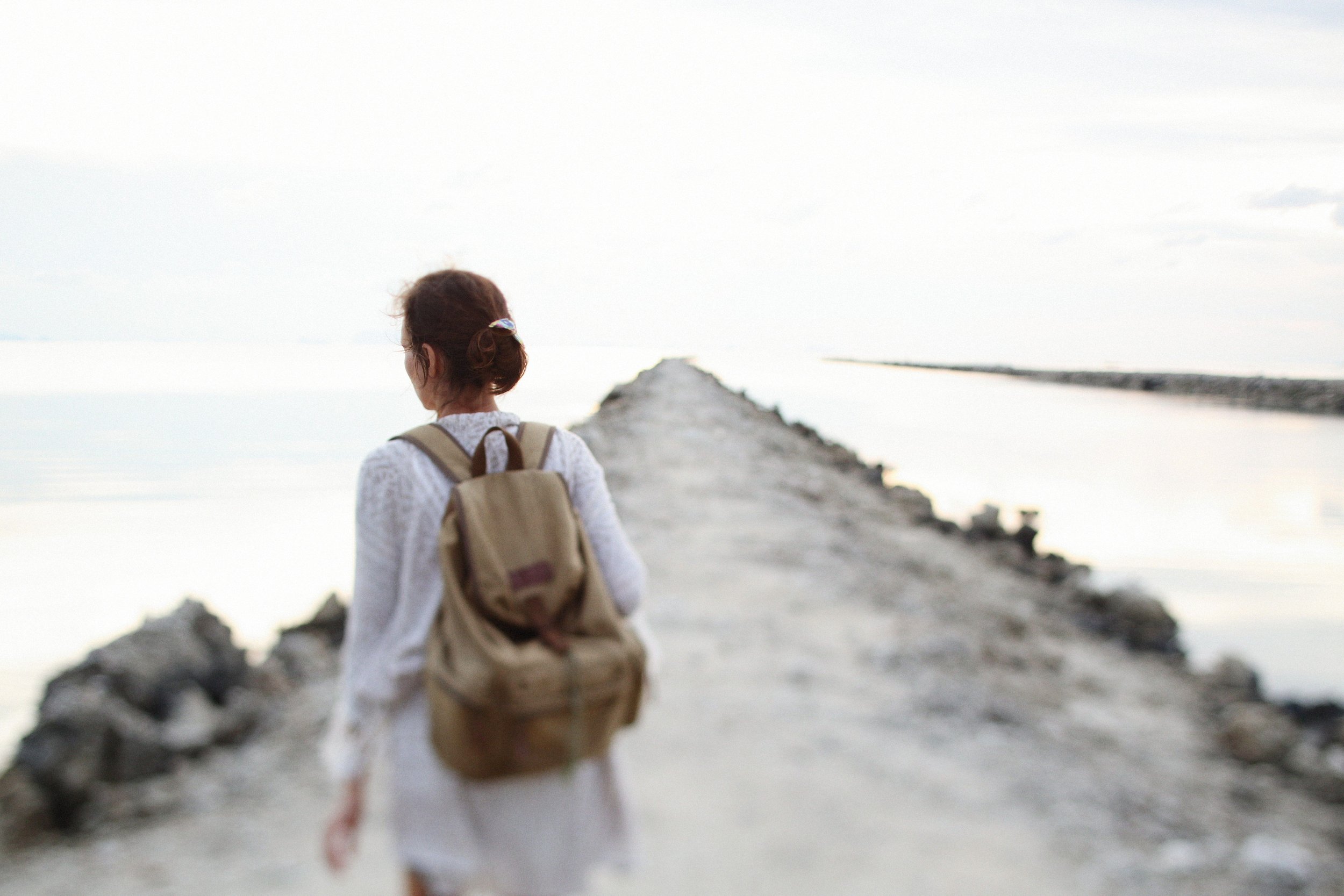 A woman with a backpack walking on a rocky pier or breakwater into a body of water, with an overcast sky.