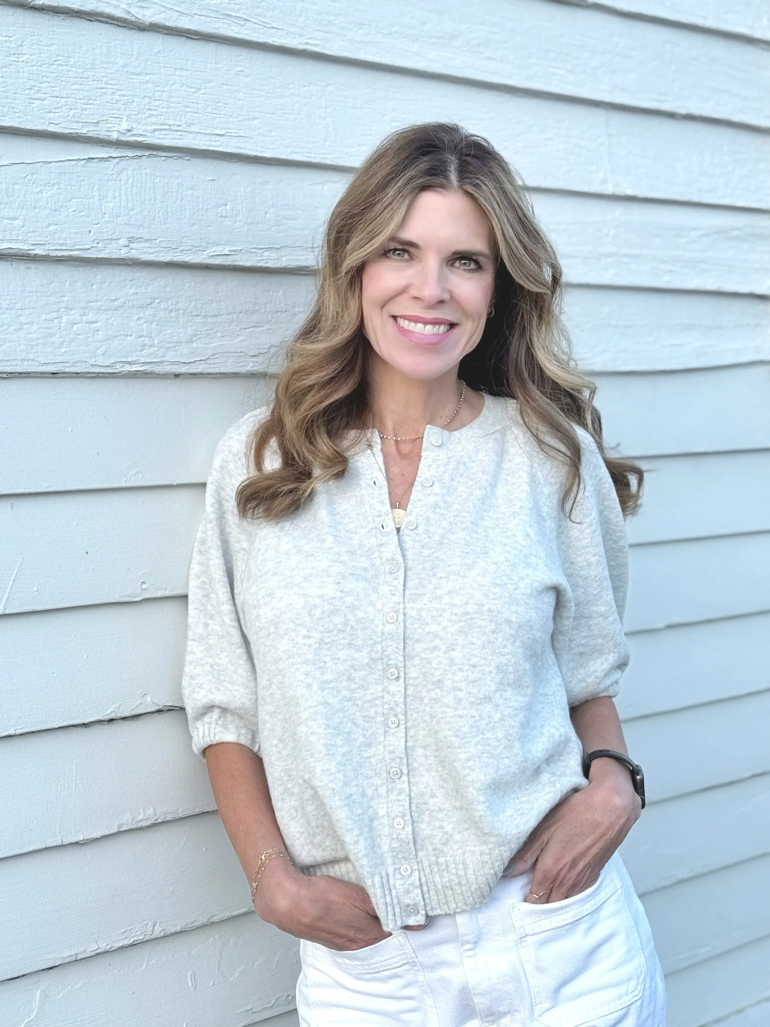 A woman with long wavy blonde hair smiling, wearing a light gray button-up top and white pants, standing against a white wooden wall.