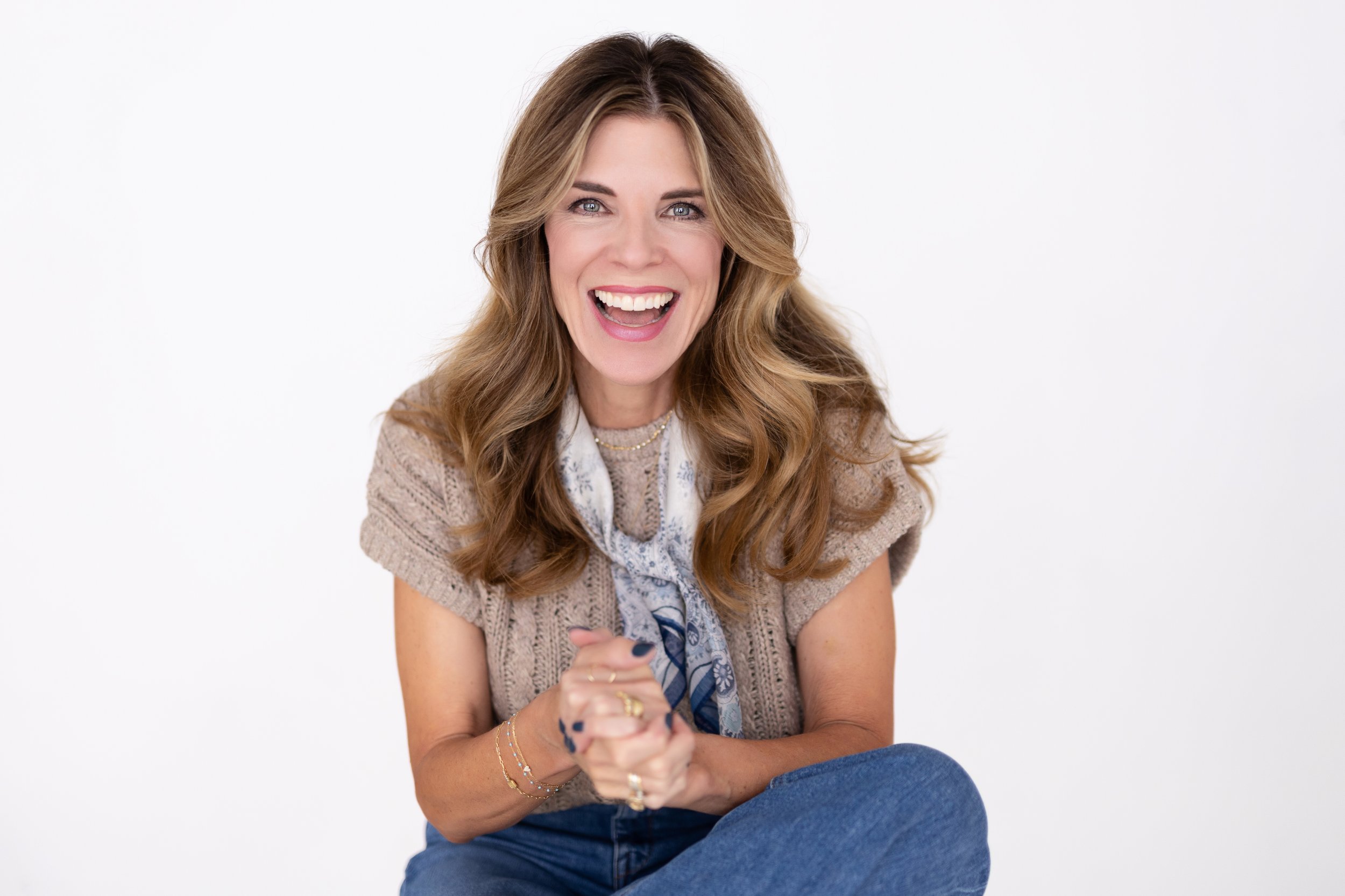 A woman with long wavy brown hair, wearing a beige knitted short-sleeve top and a blue scarf, smiling broadly with teeth showing, against a plain white background.