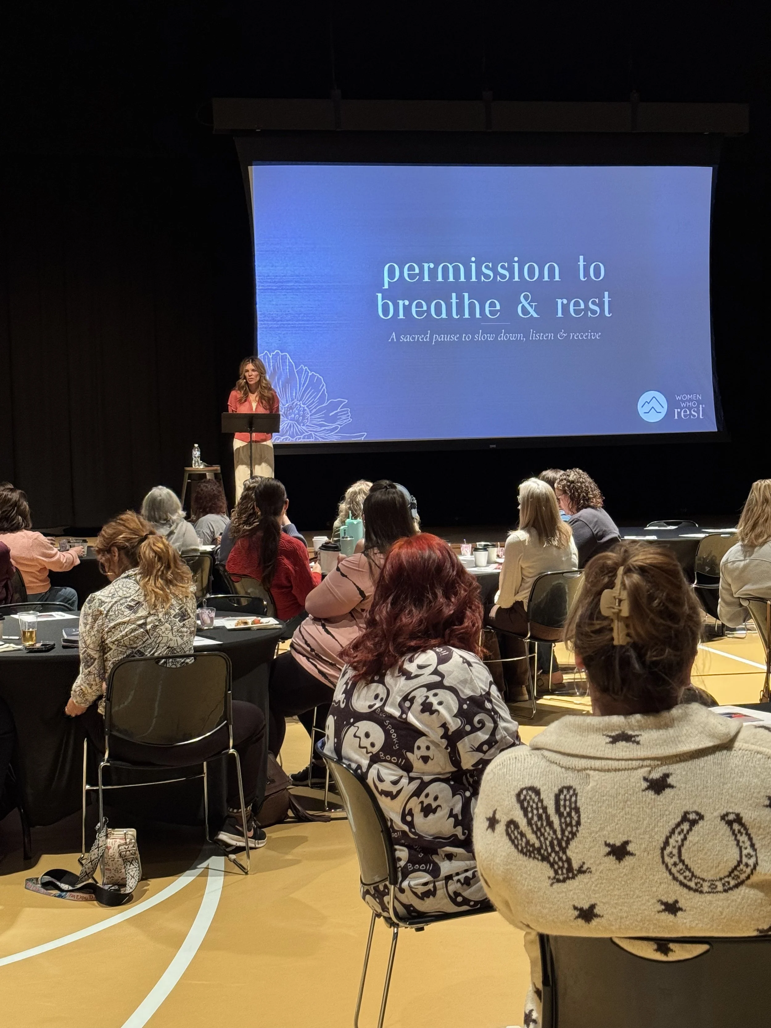A woman speaking at a conference with a large screen behind her displaying the words 'permission to breathe & rest' and the logo 'Women Who Rest'; audience members seated at tables in a gymnasium or auditorium.
