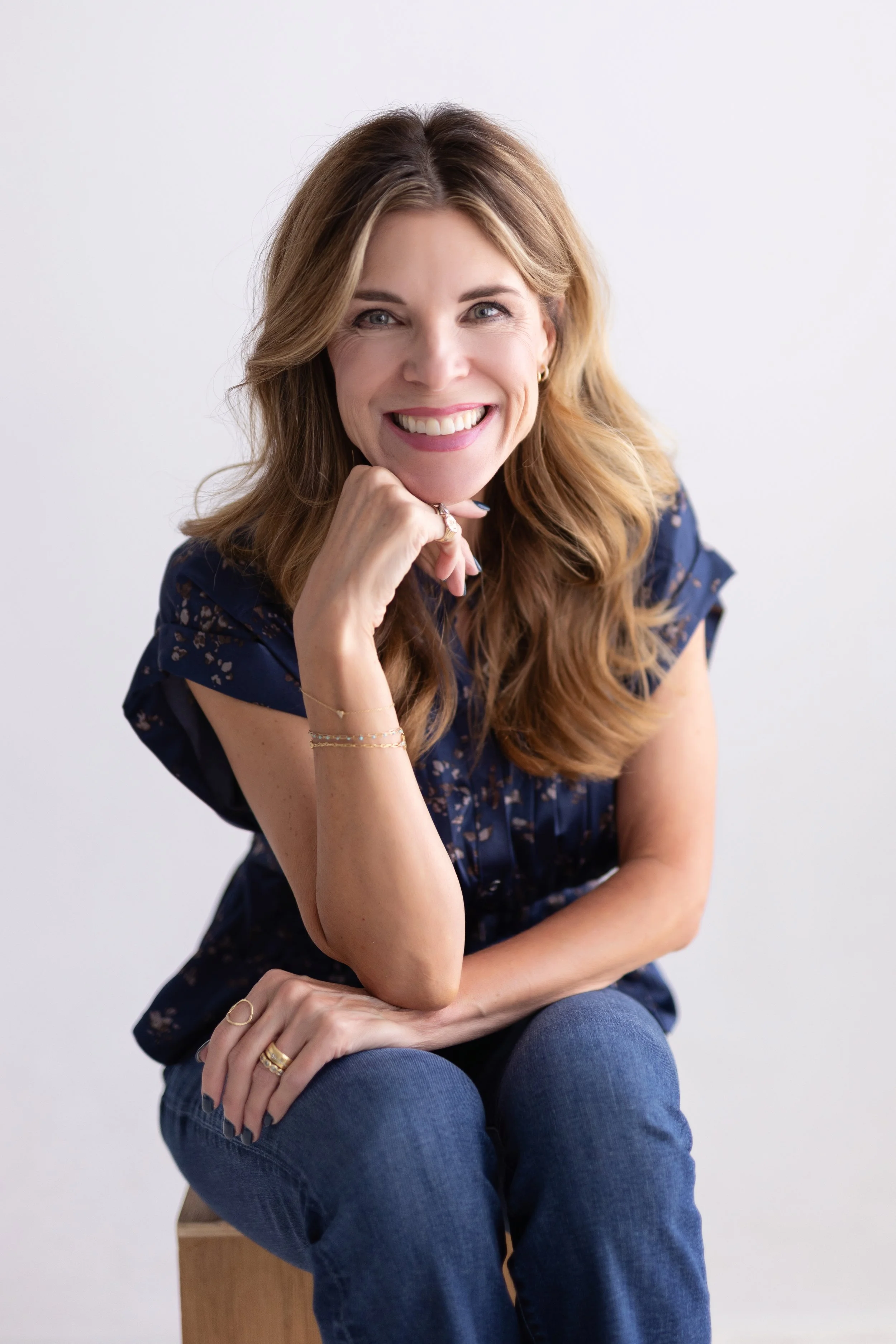 Woman with long wavy red hair smiling, sitting on a wooden stool against a plain white background, wearing a navy blue floral blouse and blue jeans.