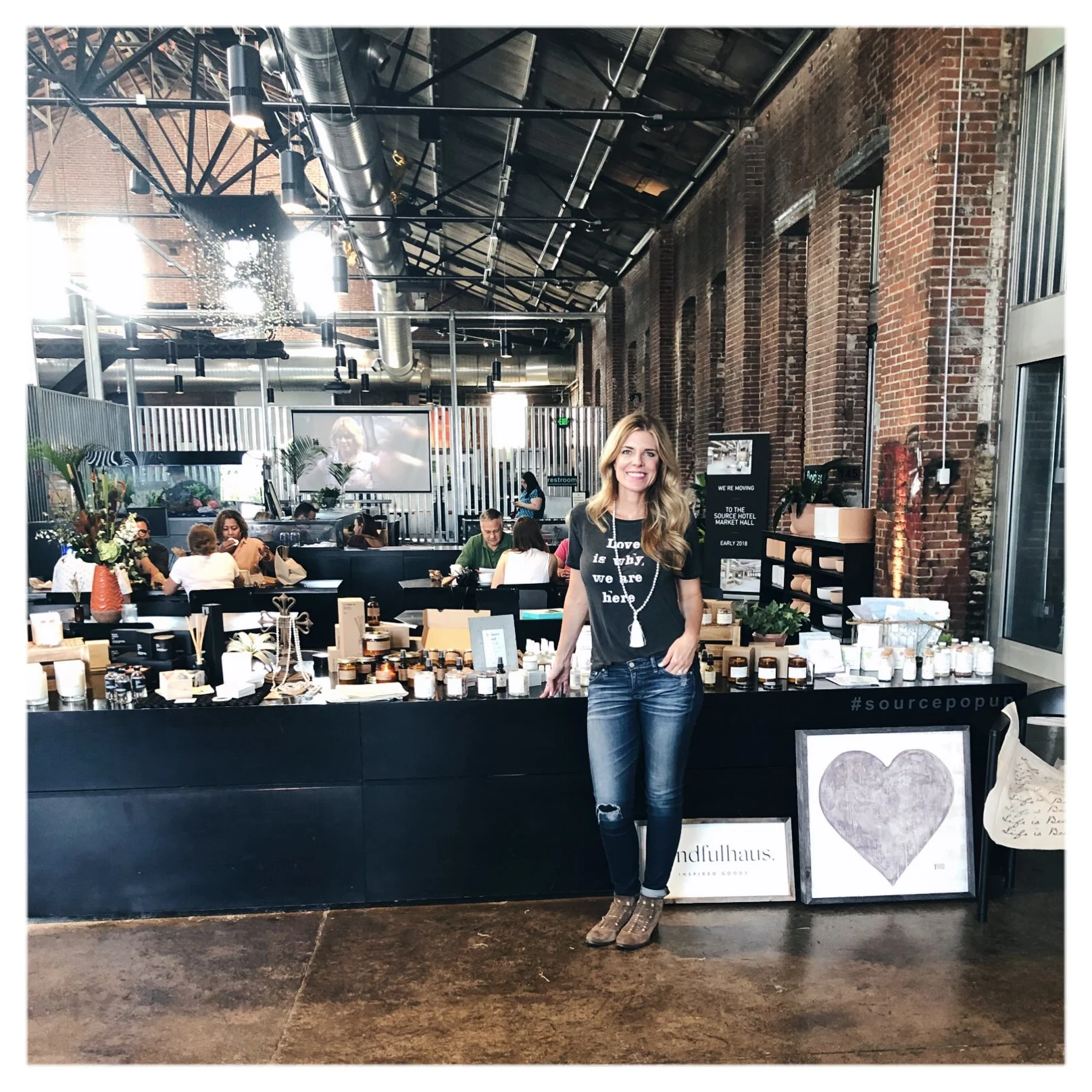 A woman standing in front of a retail display with various products such as candles and framed art. She is smiling and wearing a black t-shirt and ripped jeans in a modern, industrial-style market hall with exposed brick walls and metal ductwork.