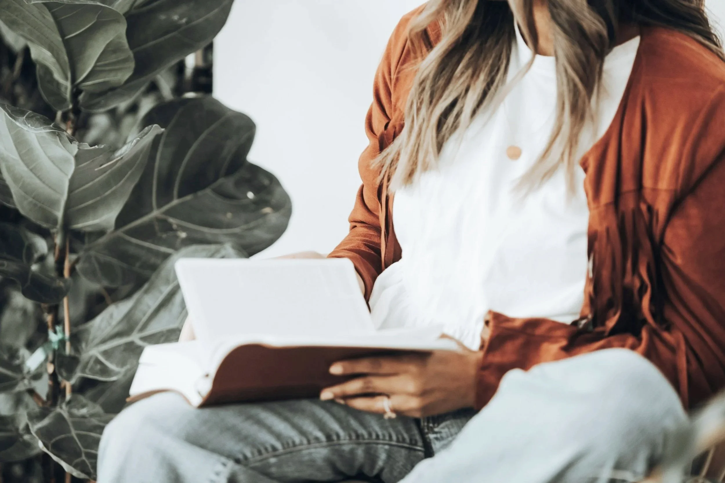 A person wearing a white shirt, orange-brown jacket, and gray pants sitting indoors near a large green leafy plant, using a tablet device.