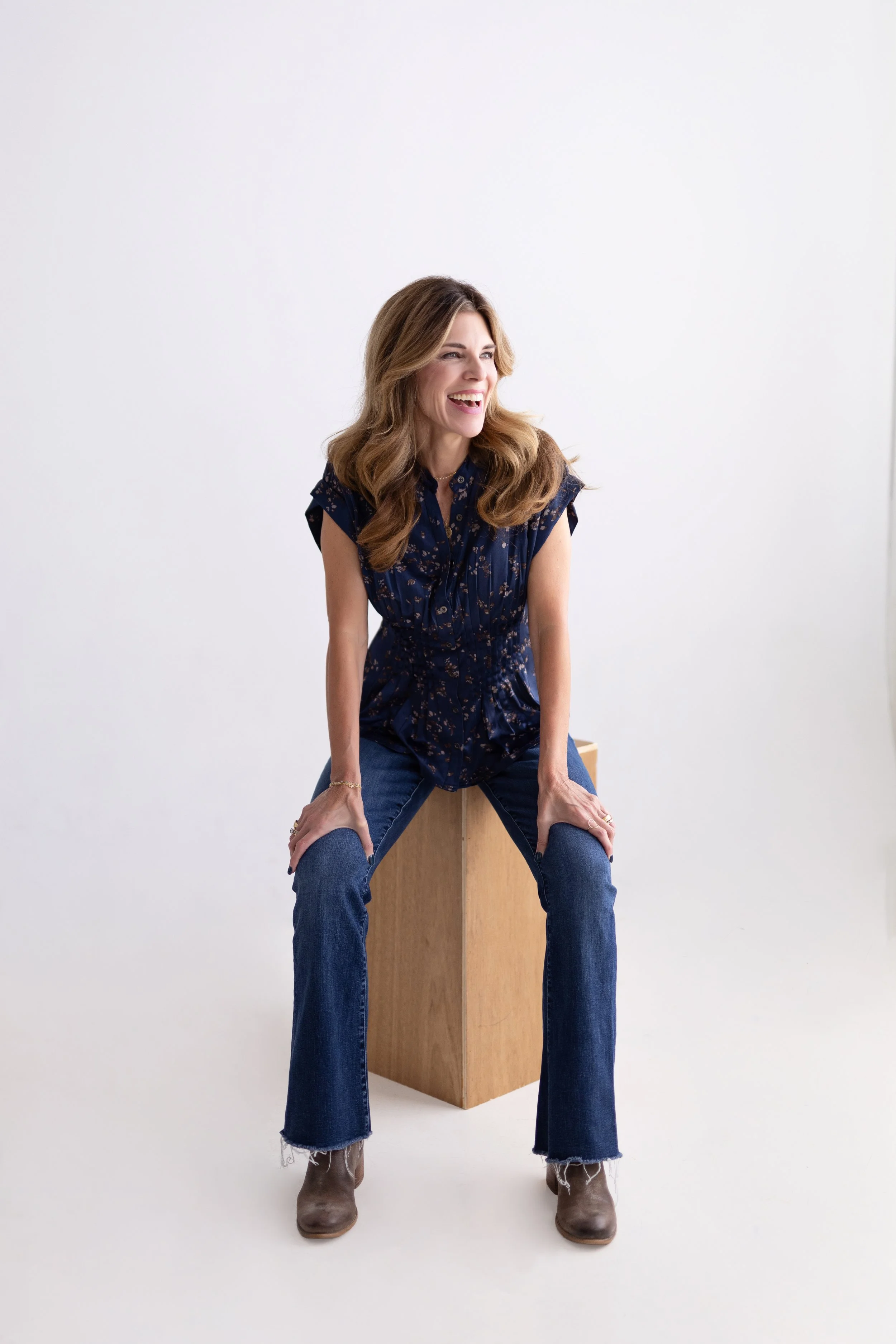 A woman with long brown hair wearing a blue patterned top, blue jeans, and brown boots, sitting on a wooden box against a plain white background, smiling and looking to her left.