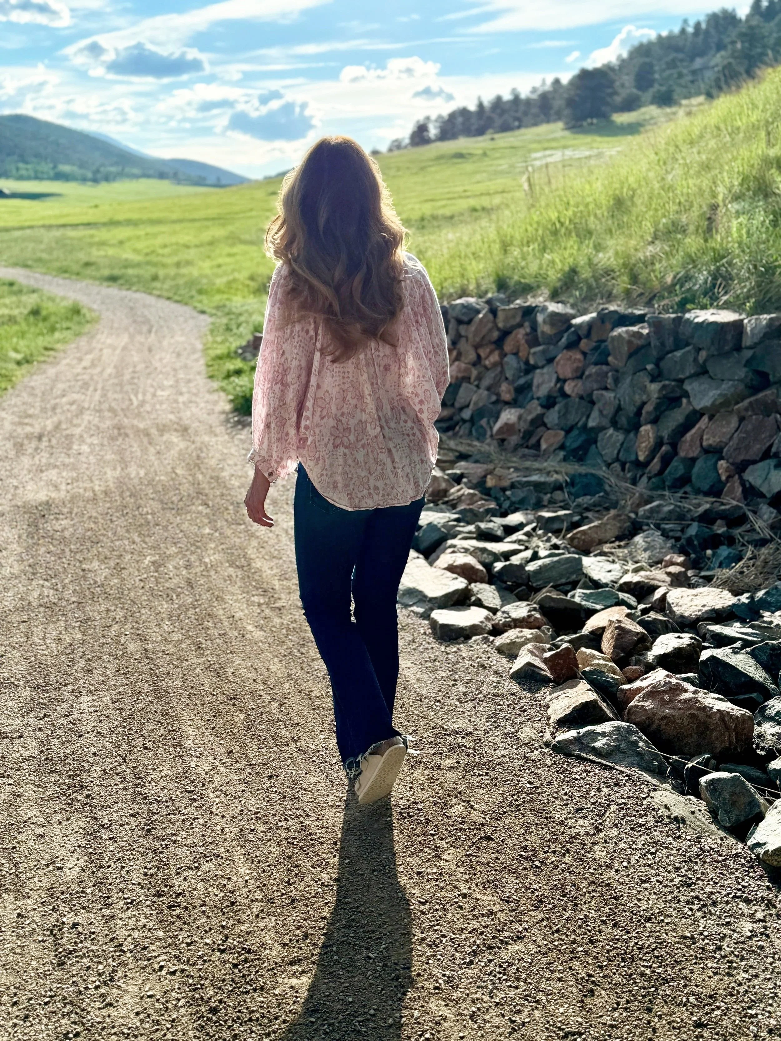 A woman with wavy hair wearing a pink patterned blouse and dark jeans walking along a gravel path beside a rocky creek in a rural landscape with green hills and a blue sky with clouds.
