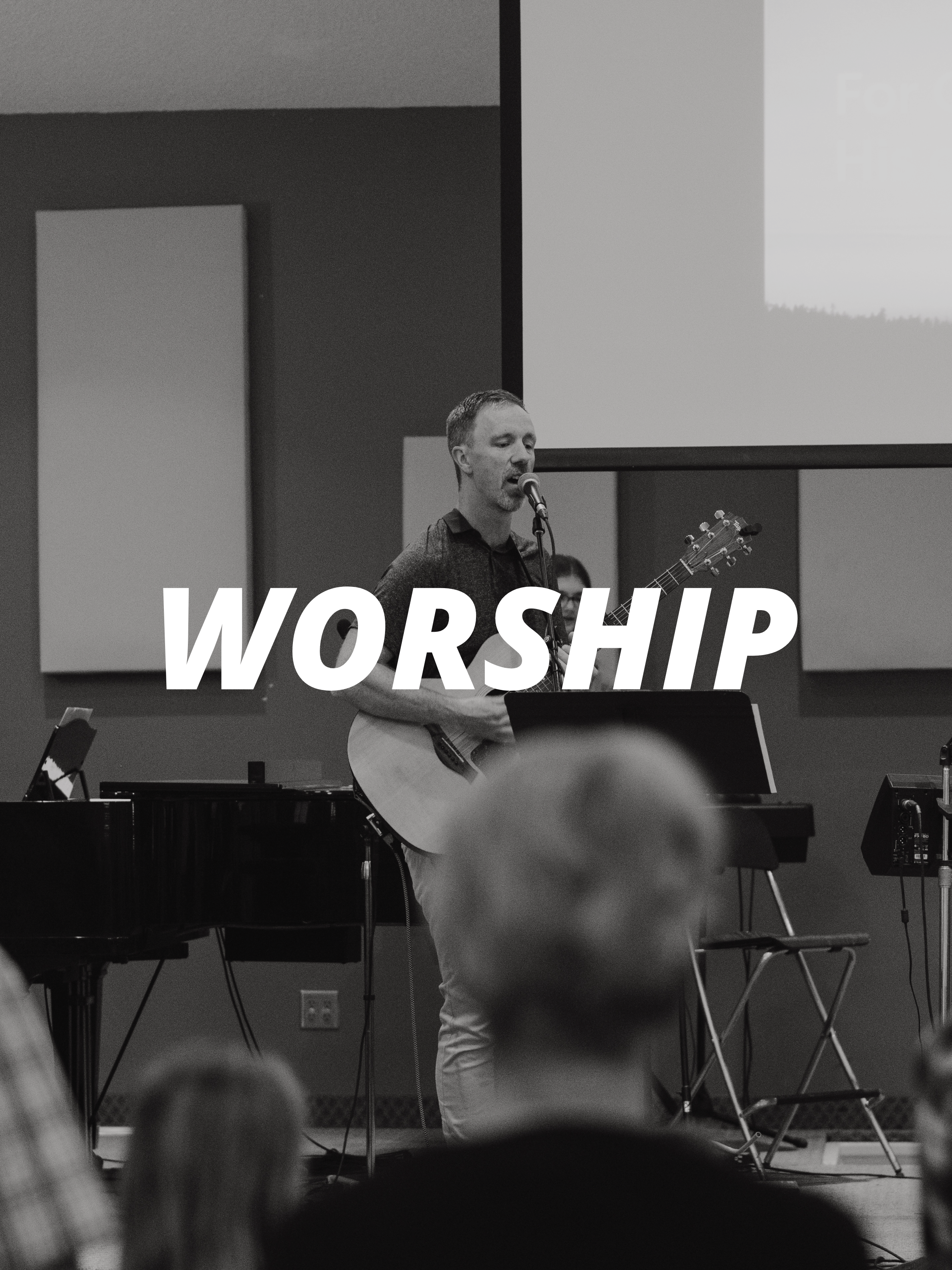 A man singing into a microphone and playing an acoustic guitar during a worship service in a church or similar setting, with a large screen in the background and several musical instruments around him.