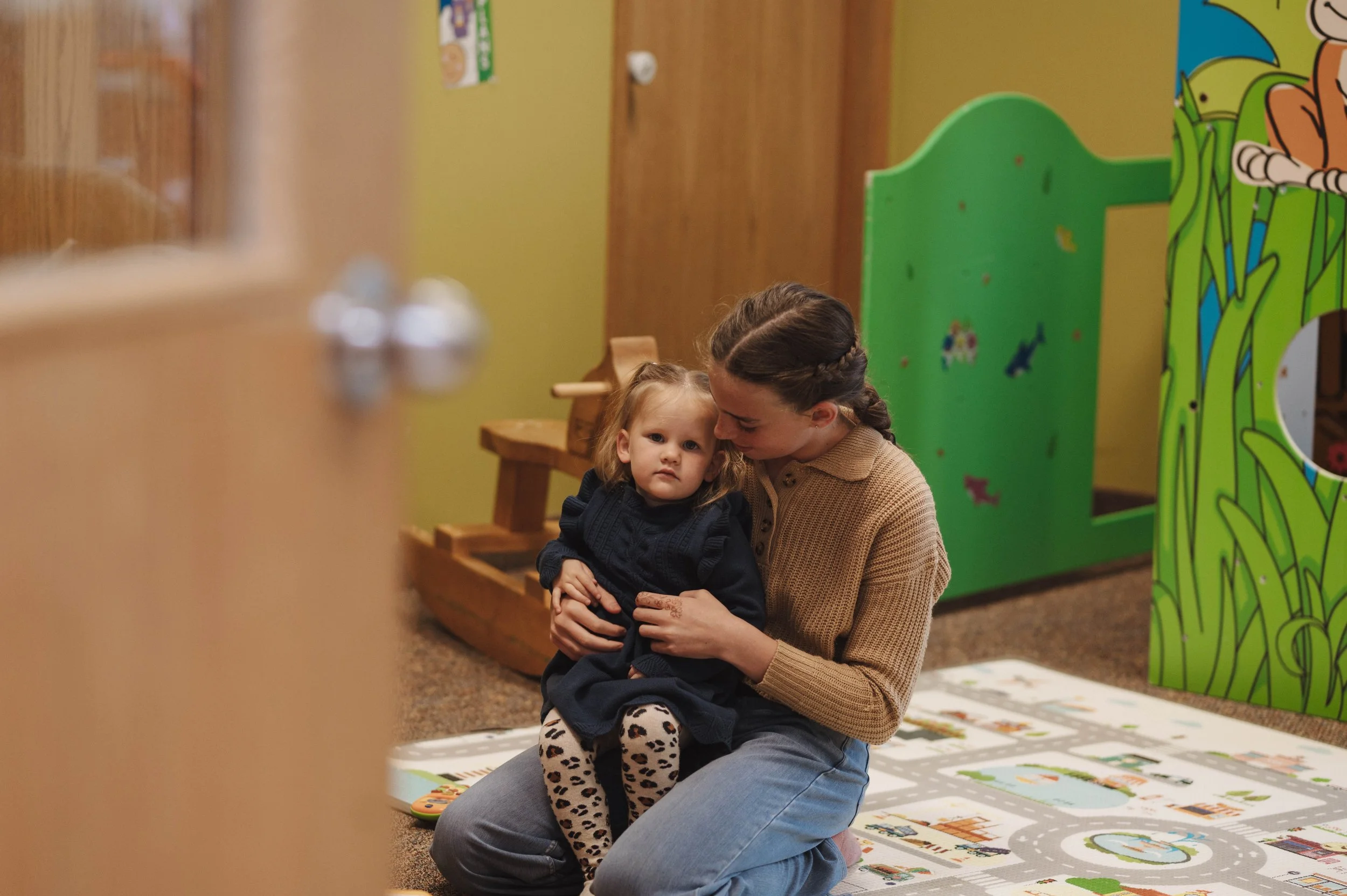 A woman with braided hair sitting on the floor, holding a young girl with blond hair and leopard print pants, both looking at the camera, in a room with colorful decorations and a green cardboard cutout.