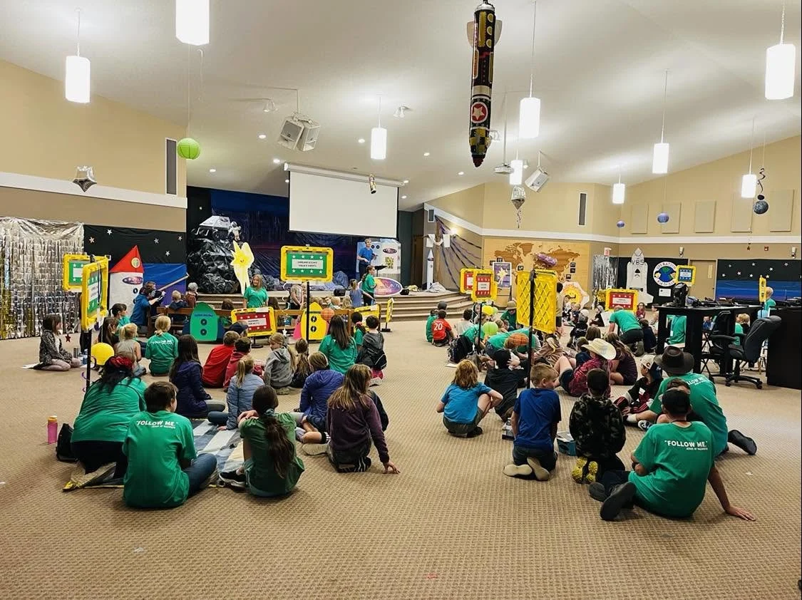 Children and adults seated on the floor in a room with a stage, playing games, possibly during a children's program or event.