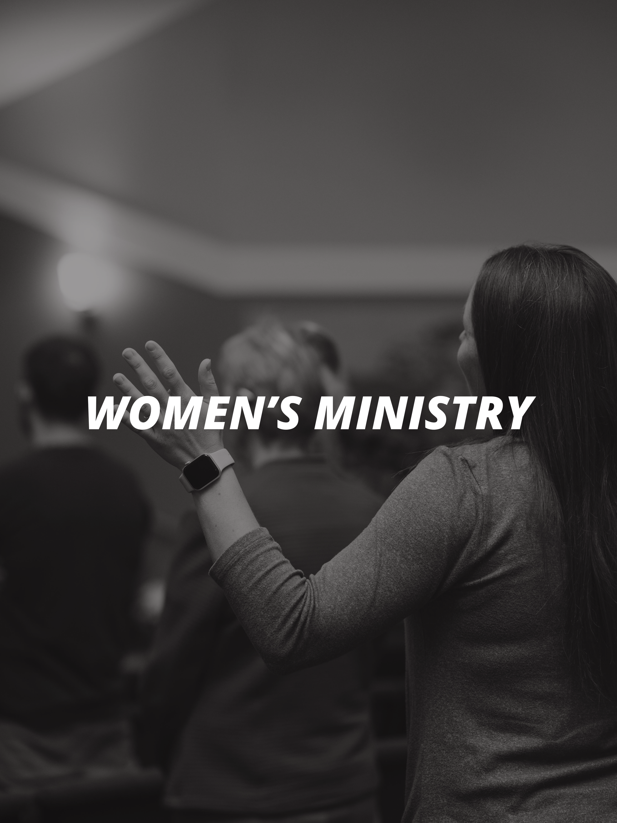 A woman with long hair wearing a smartwatch on her left wrist, raising her right hand in a prayer or worship gesture at a women's ministry event.