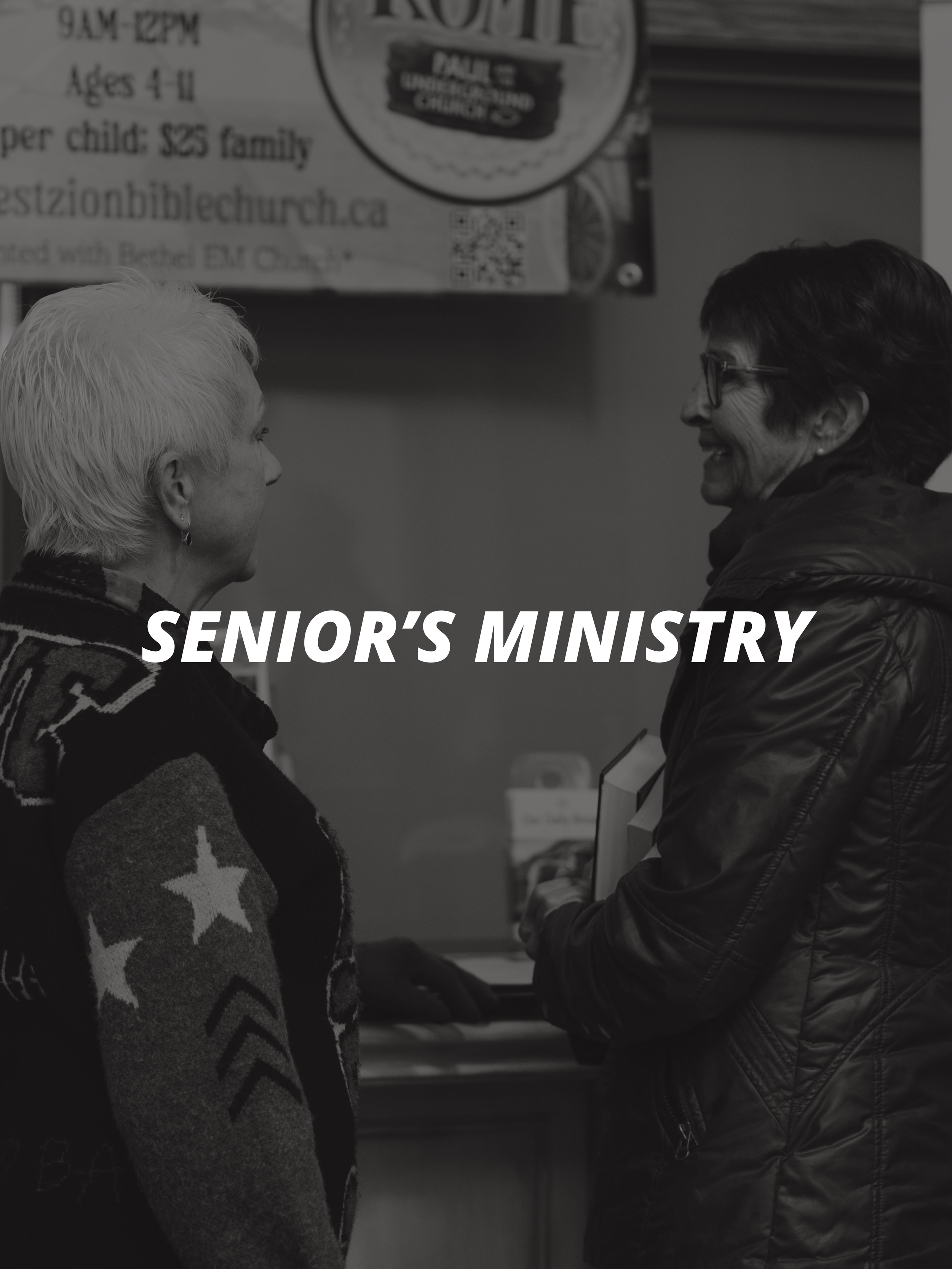 Two women talking, one with short white hair and patterned sweater, the other with glasses and dark coat, inside a church or community center, with a banner overhead reading 'Senior's Ministry'.
