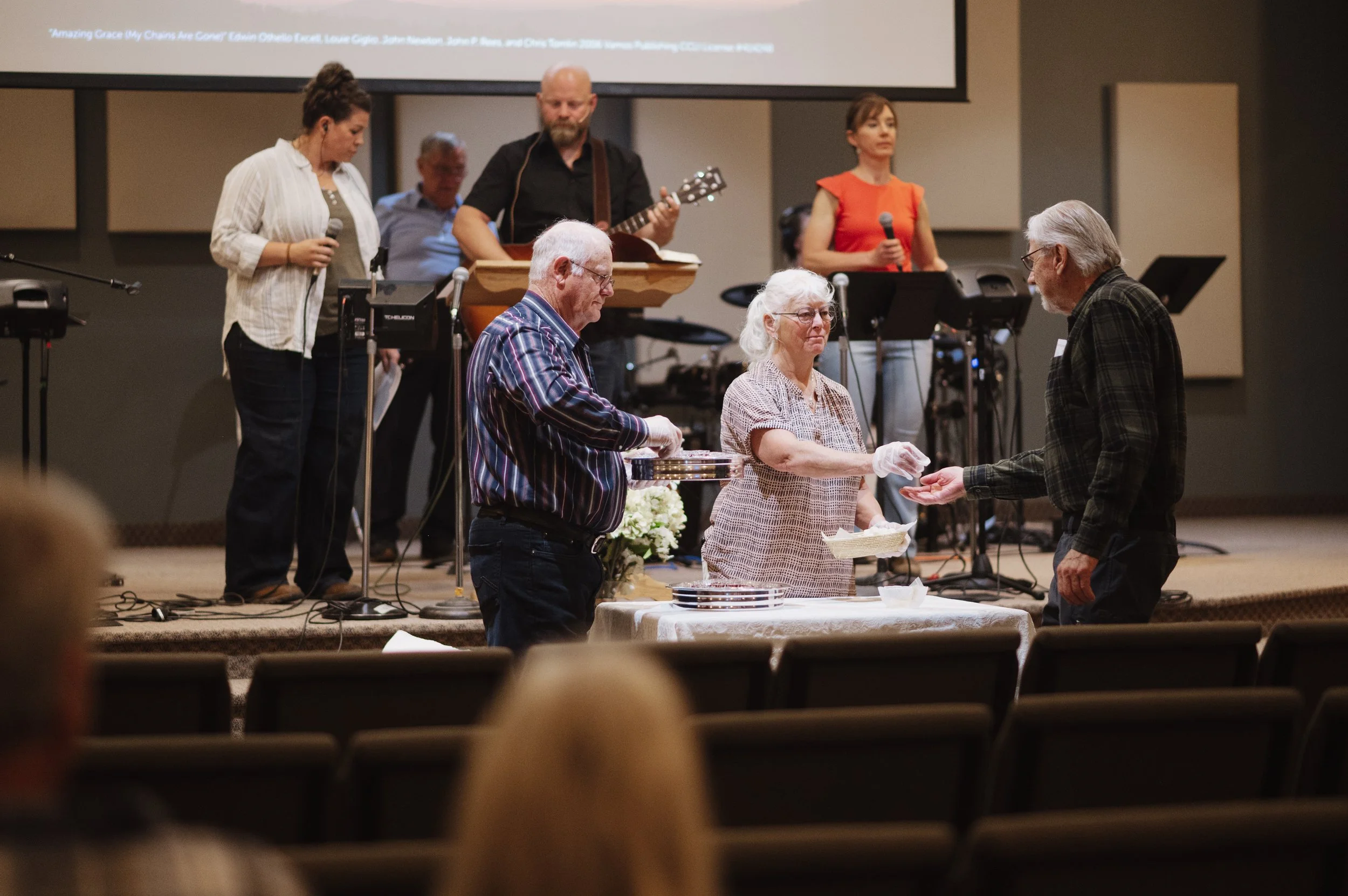 A church or community hall setting with a group of elderly people at an event. Three individuals are at the front: one man and two women, involved with serving or receiving a meal or donation, while a band is performing in the background, including a man playing guitar. Two women in the background are holding microphones, possibly preparing to sing.