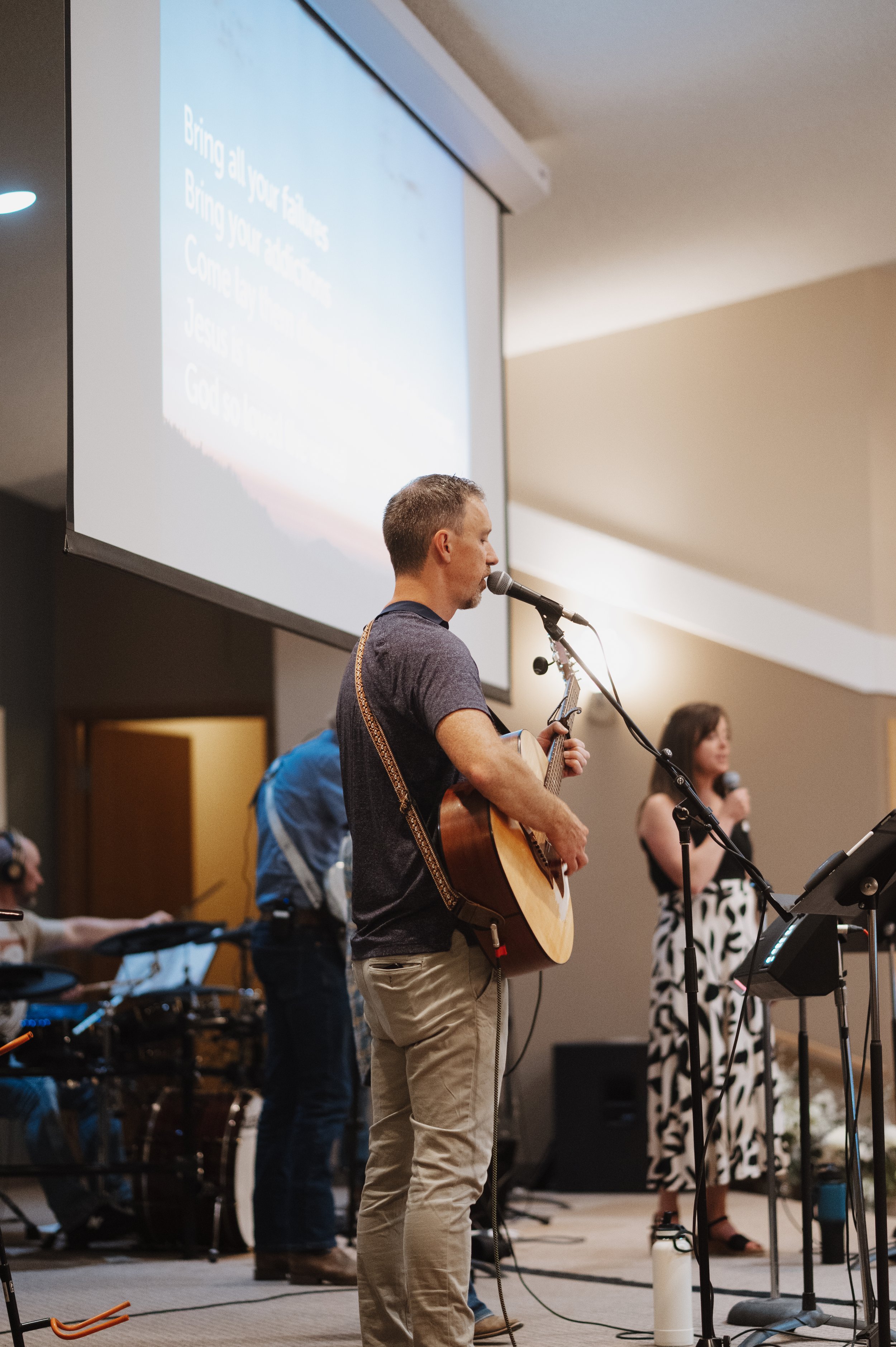 A man playing guitar and singing into a microphone during a church service or worship session, with a woman singing in the background and other musicians and equipment around at Bethel Church near Carstairs AB