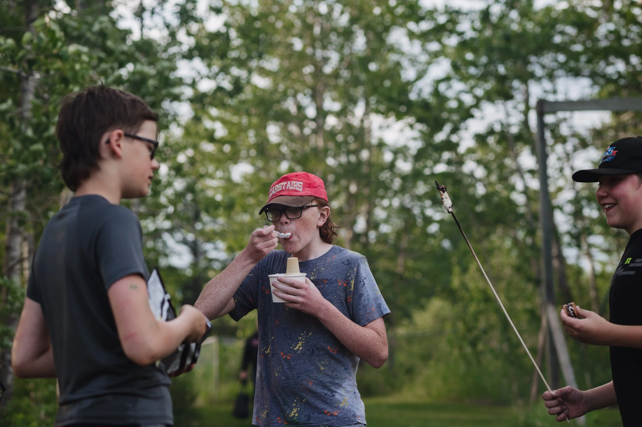 Group of young friends outdoors, one eating ice cream, others smiling and talking, in a lush green park at Bethel Church youth event near Carstairs AB.