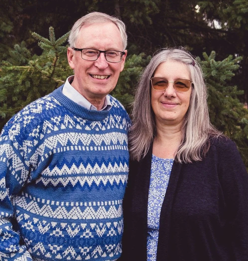 A smiling man and woman standing outdoors in front of a pine tree, dressed in casual winter clothing.