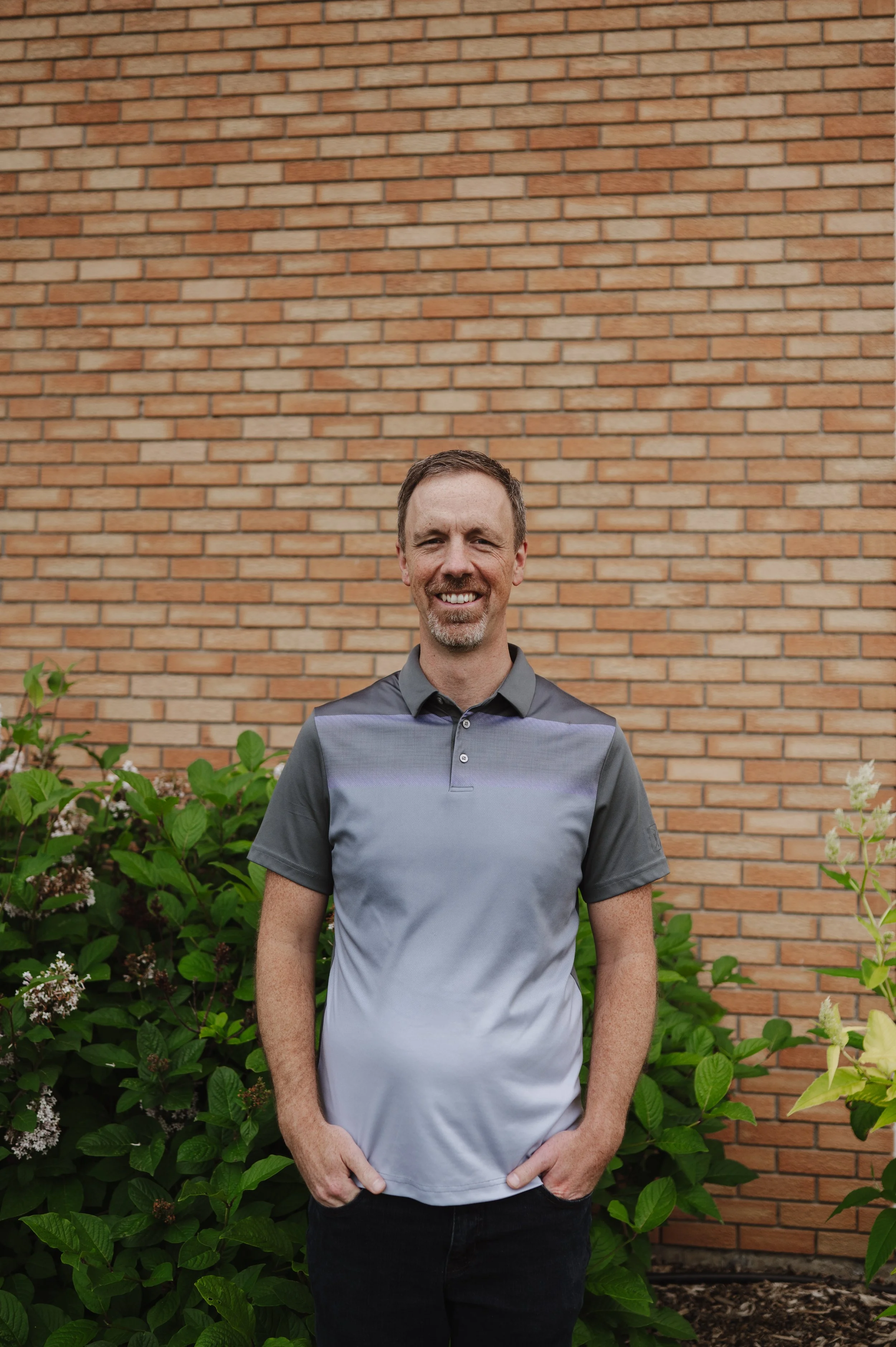 A man with short brown hair, a goatee, and a big smile standing outdoors in front of a brick wall and green plants.
