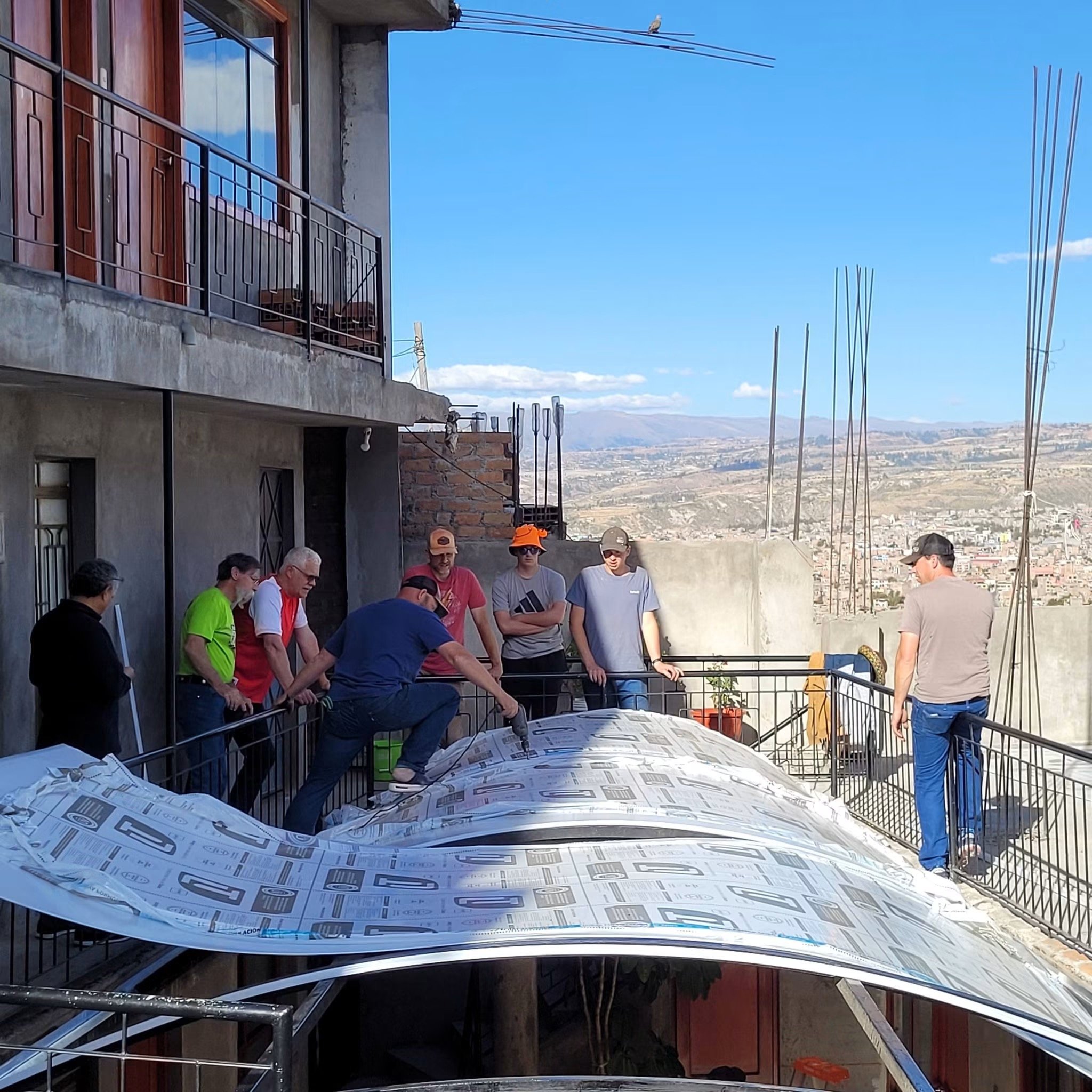 A group of people standing outdoors on a sunny day, some wearing hats and sunglasses, in front of a building with a balcony and large windows. Construction materials and pipes are visible, indicating ongoing construction or renovation work.