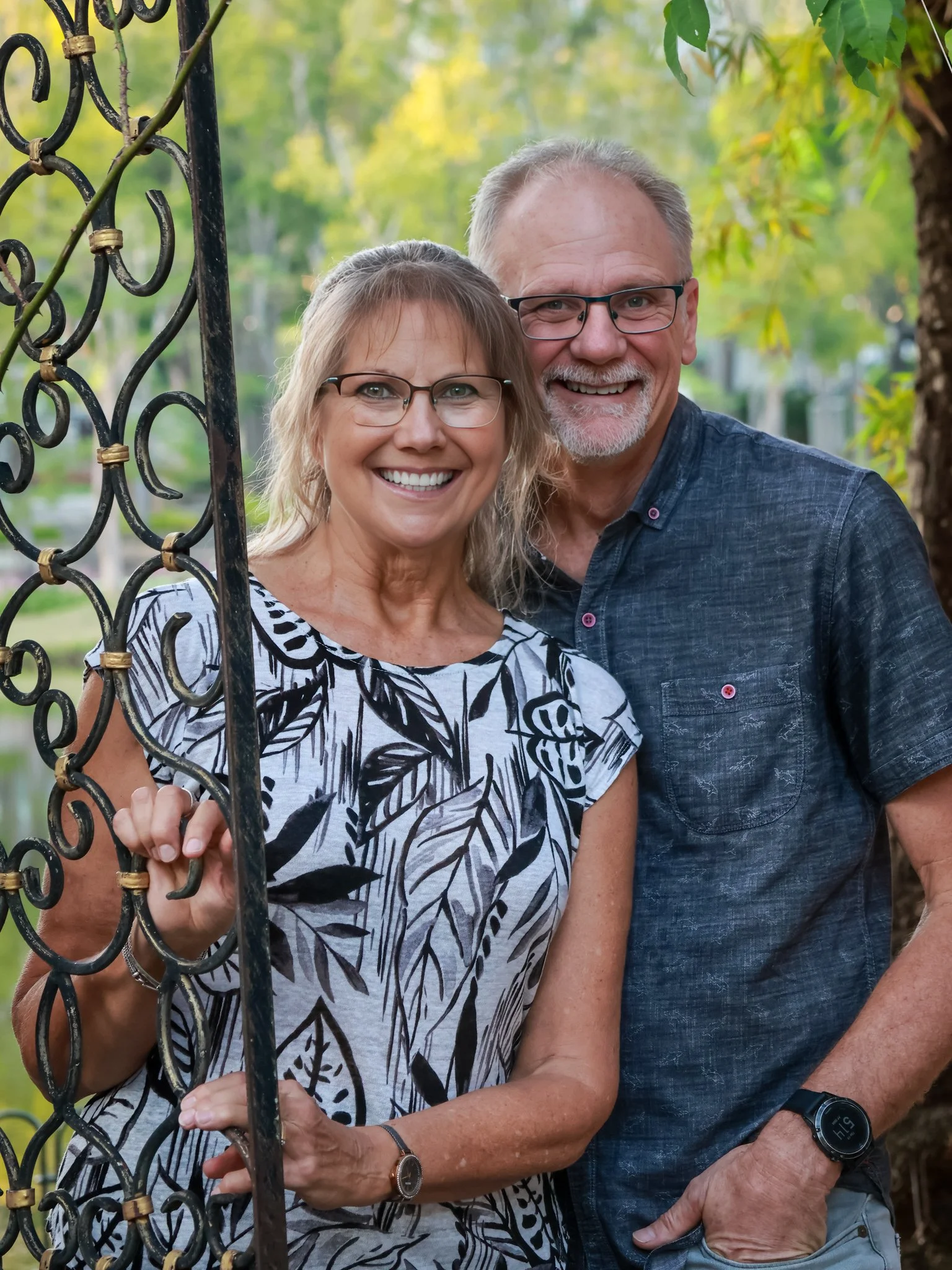 A smiling middle-aged woman and a smiling middle-aged man standing outdoors near a black decorative metal fence, surrounded by green trees and foliage, during daytime.
