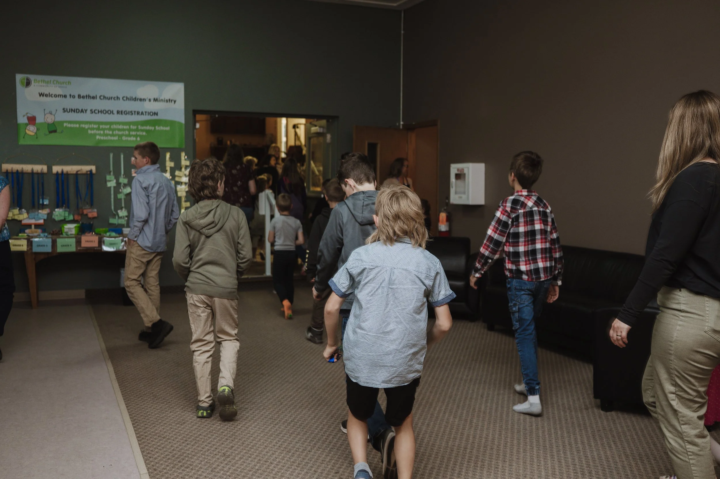 Children and adults walking in line inside Bethel church, with a sign for Sunday school registration visible on the wall.