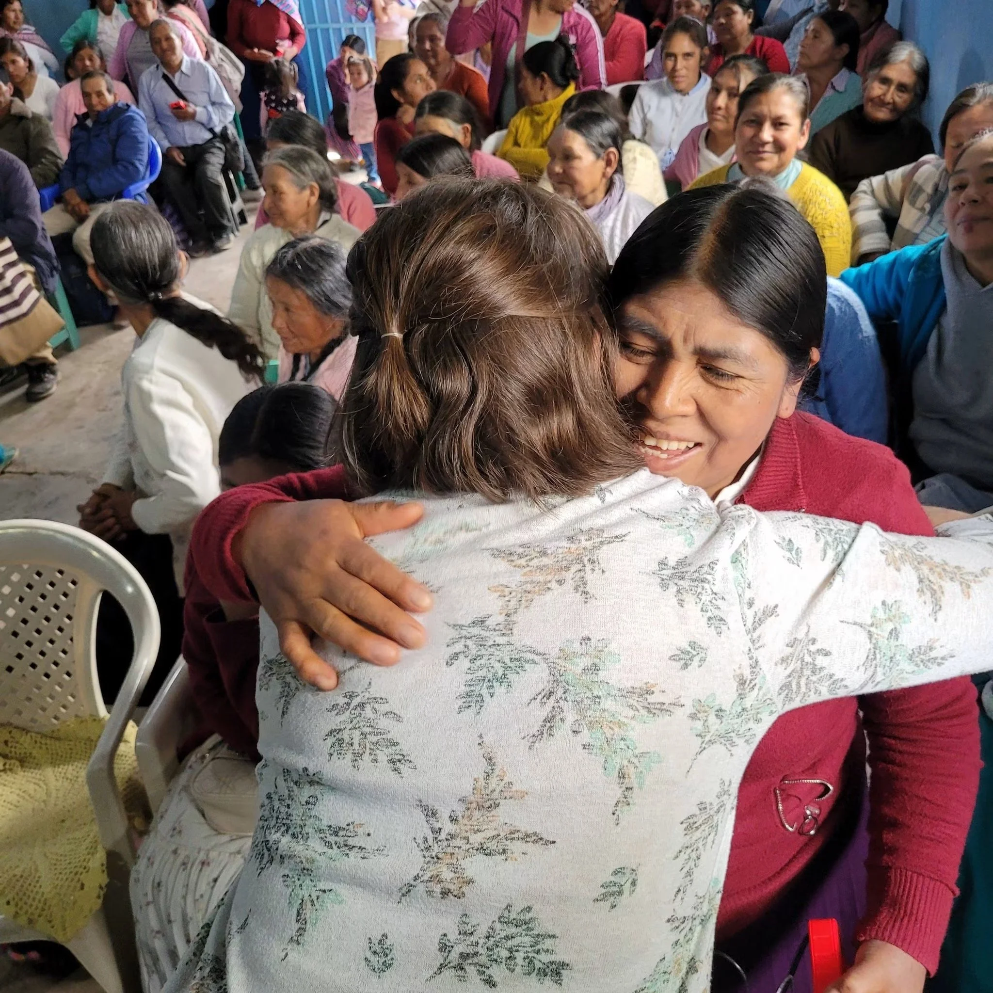 A large group of women and some children gathered together, with some standing and some sitting, sharing a moment of embrace and connection in an indoor setting with blue walls.