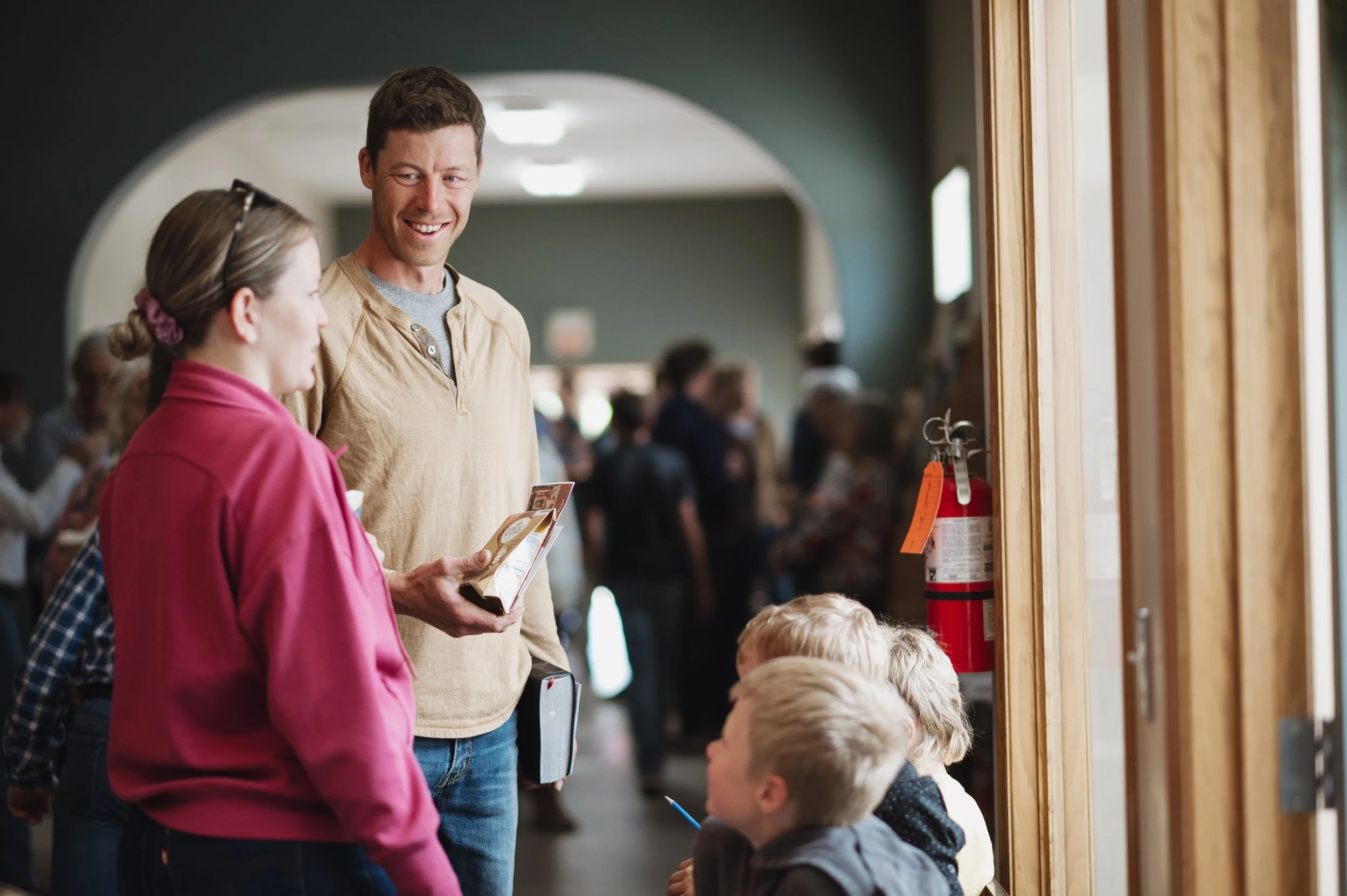 A man and woman are talking to children in a crowded indoor setting, after the Sunday morning service at Bethel Church near Carstairs AB.