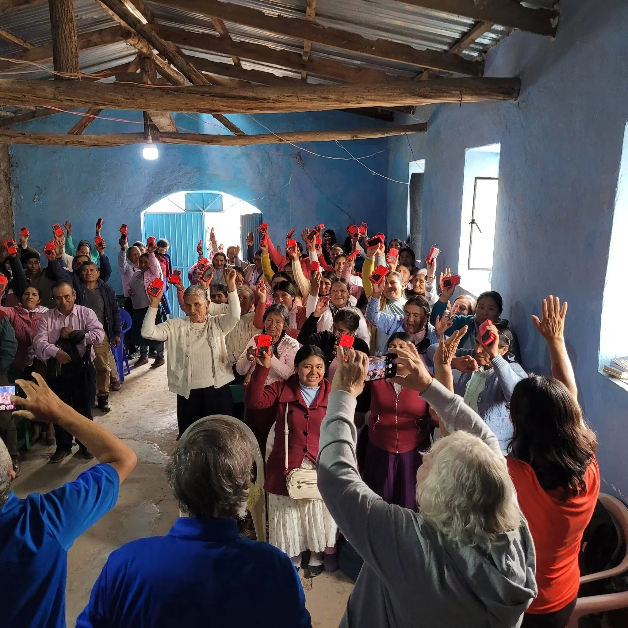 A group of people gathered indoors holding up red and black devices, with a rustic wooden ceiling and bright natural light coming from a window or door in the background.