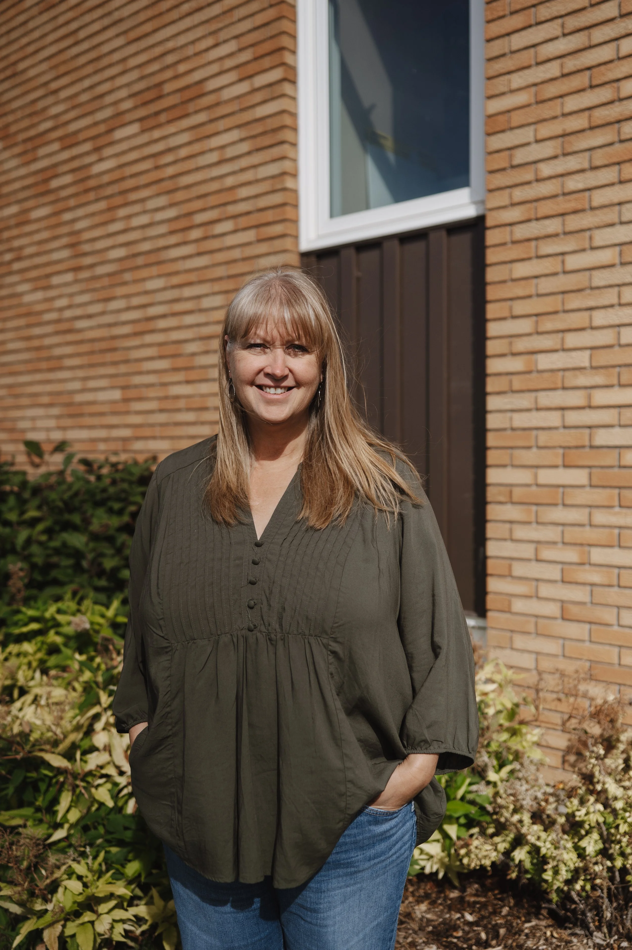 A woman with light brown hair and bangs, wearing a white sweater and a brown patterned jacket, standing outdoors in front of a brick wall and green foliage.