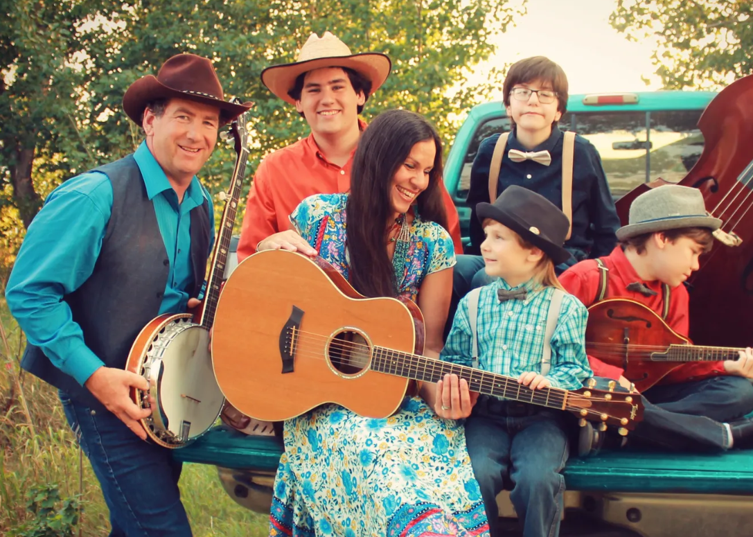 A family of six with light skin and three children, wearing vintage Western and country attire, posing outdoors near a teal pickup truck with trees in the background. The mother is playing an acoustic guitar, and others are holding musical instruments or sitting on the truck bed, smiling and enjoying a sunny day.
