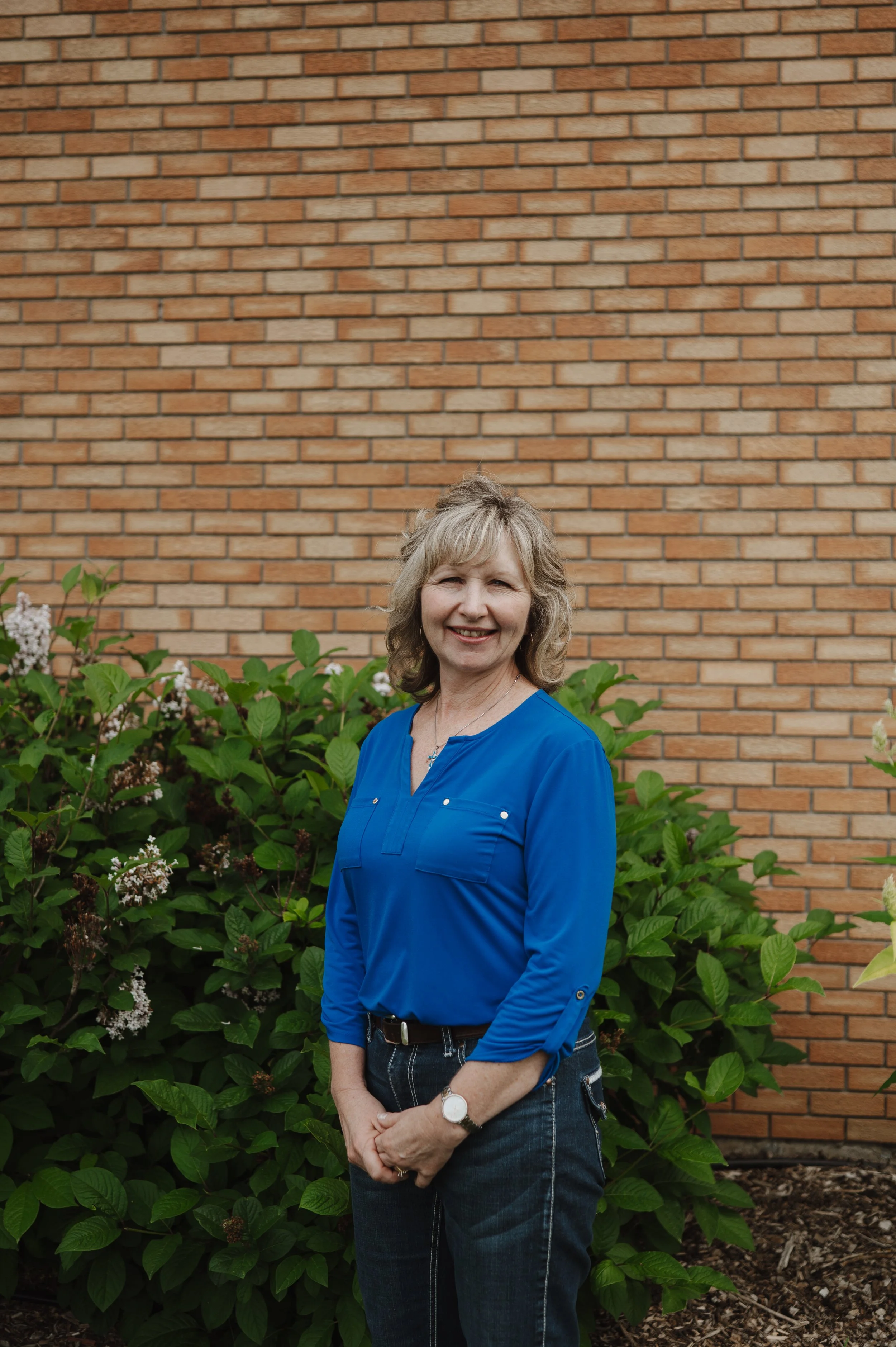 A middle-aged woman with blonde hair, wearing a bright blue blouse and dark jeans, standing outdoors in front of green bushes with white and pink flowers, and a brick wall in the background, smiling at the camera.
