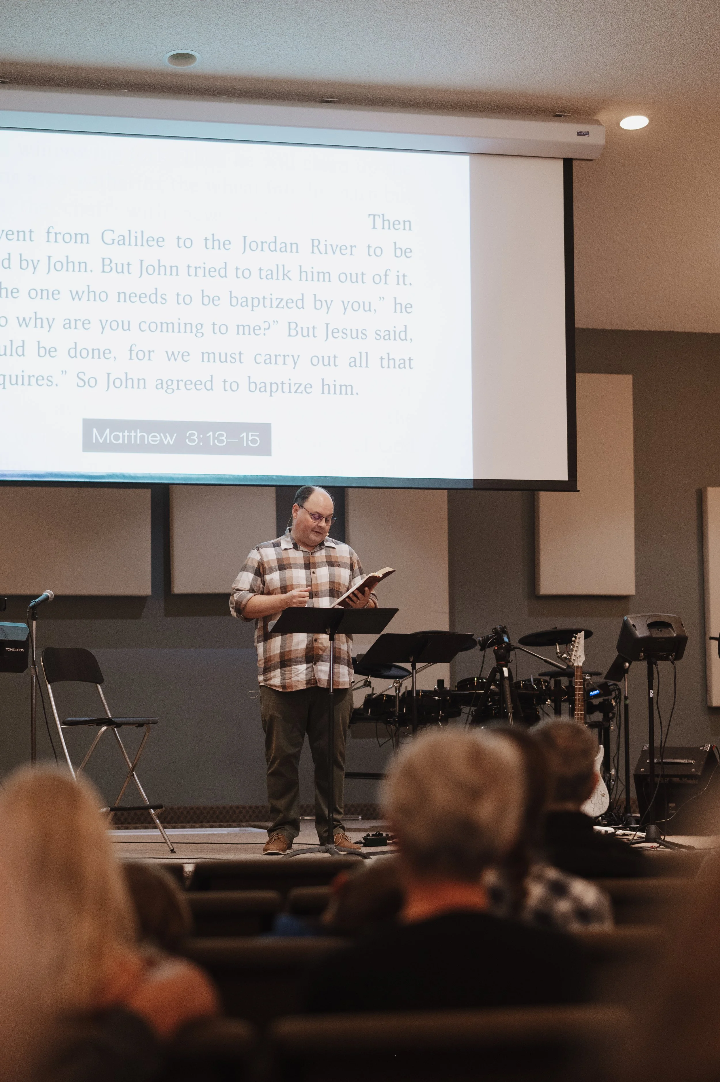 A man in glasses and a plaid shirt stands on a stage, reading from a book, with musical instruments and microphones behind him, while a woman or man appears on a large screen with biblical text and a reference to Matthew 3:13-15.
