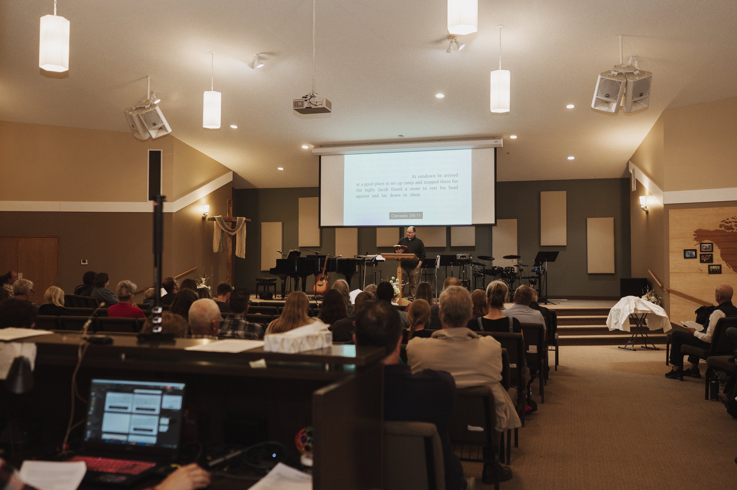 The Bethel Church auditorium with people seated, a speaker at a podium, and a large screen displaying a Bible verse from Genesis 28:11. The setting includes musical instruments and microphones, with ceiling lights and speakers.